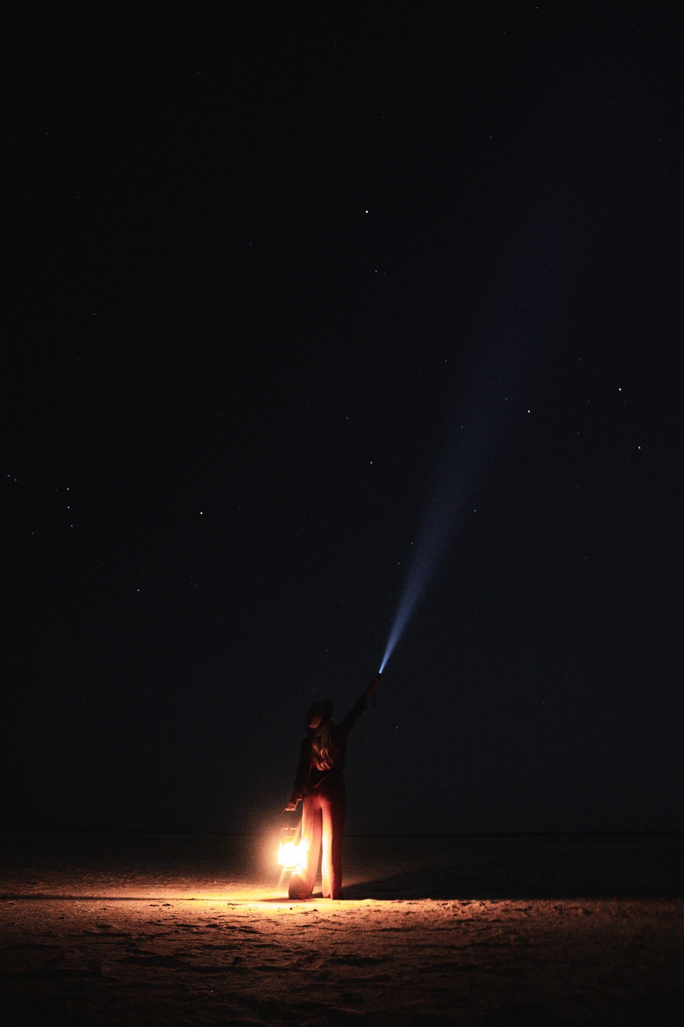 A flashlight beam cuts through the dark night sky while a lantern glows on the ground.