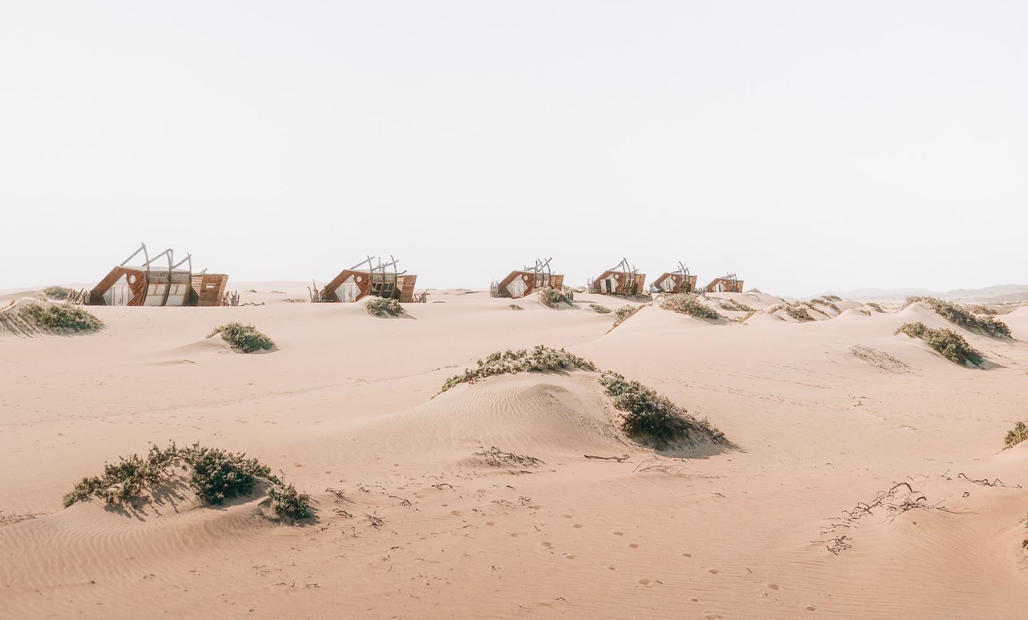 Minimalist desert landscape with scattered buildings among rolling sand dunes under overcast sky.