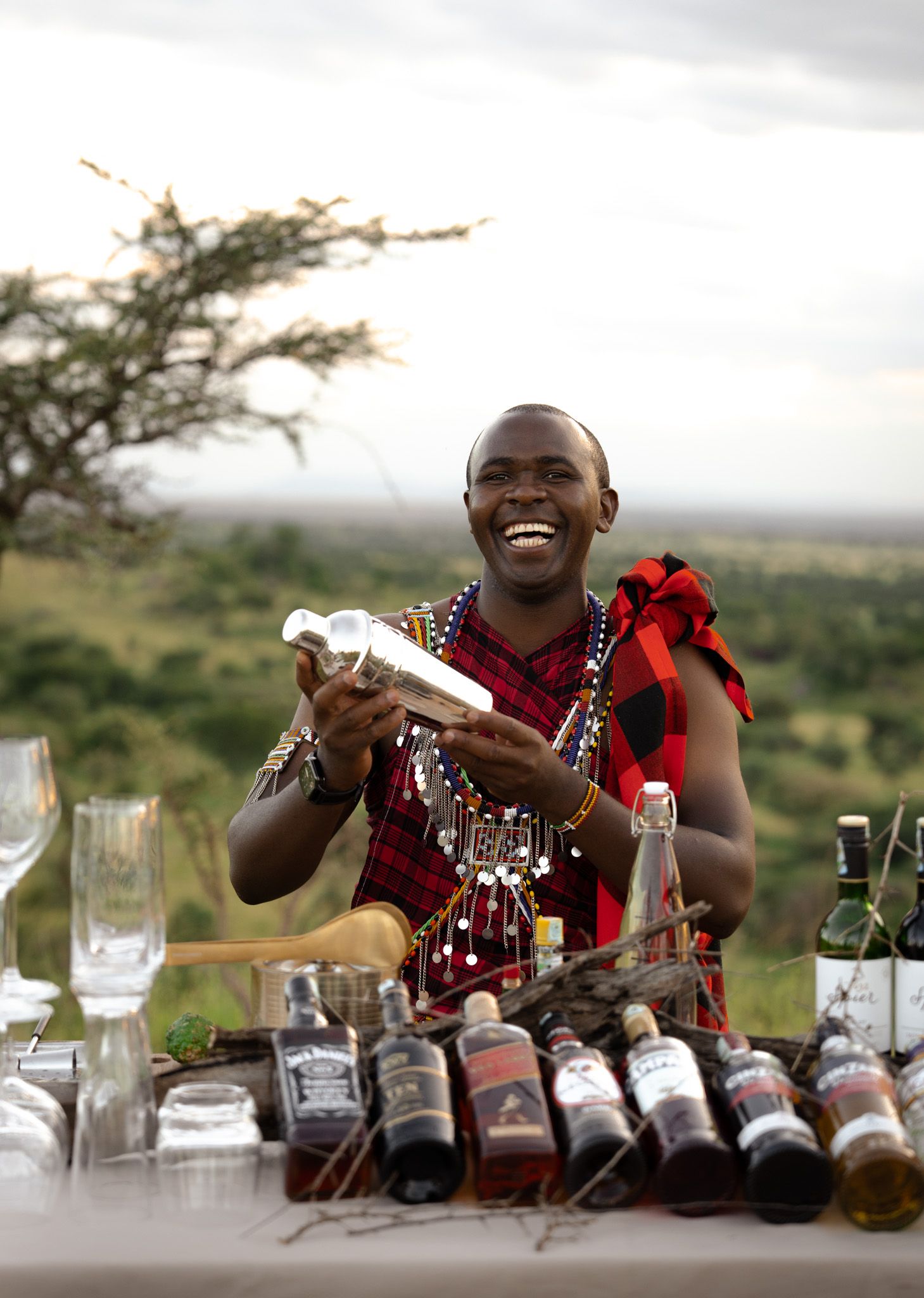 Bartender wearing traditional Maasai attire smiles while arranging bottles and glassware on an outdoor table in Kenya.