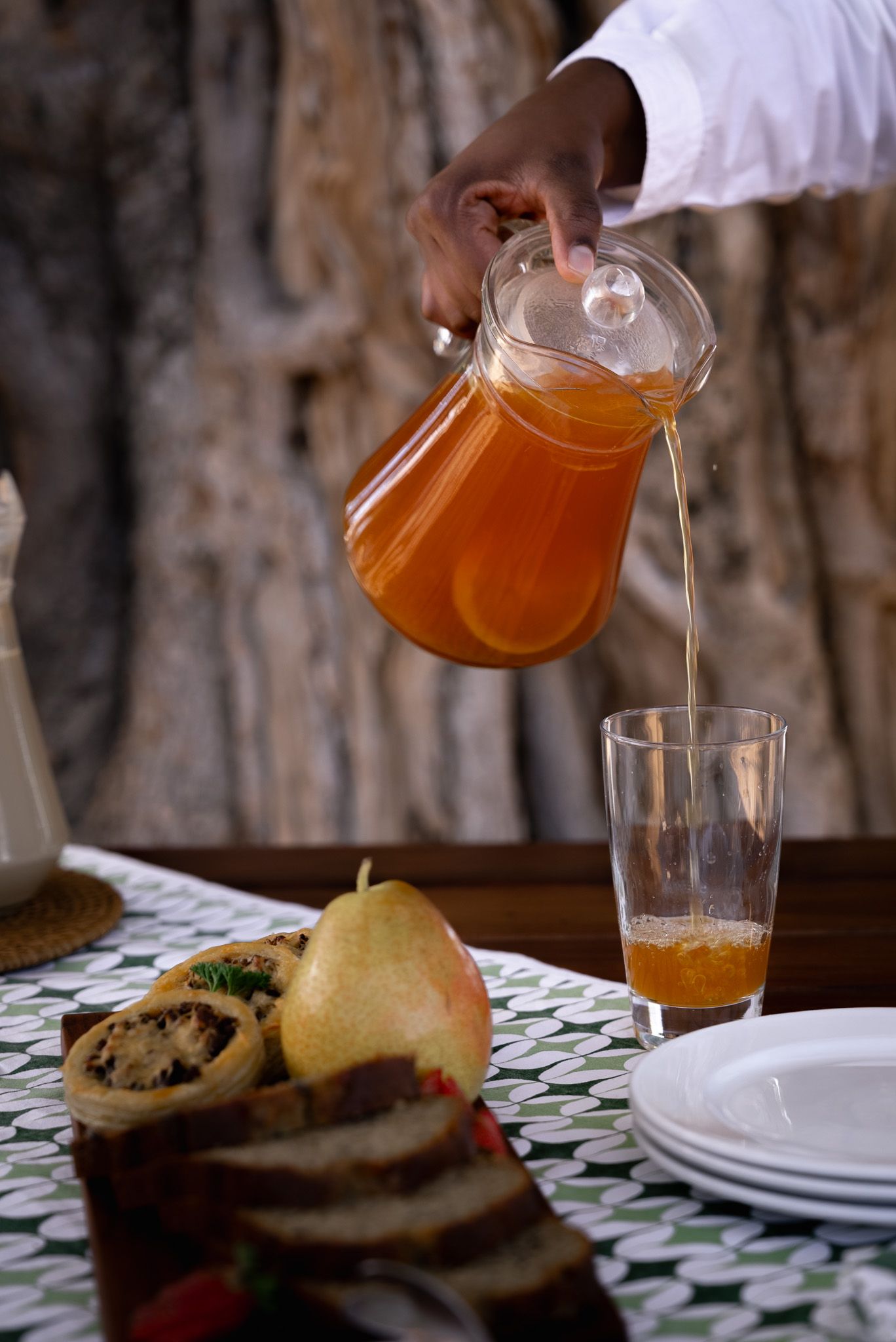 Orange drink being poured from glass pitcher into serving glass on patterned tablecloth.