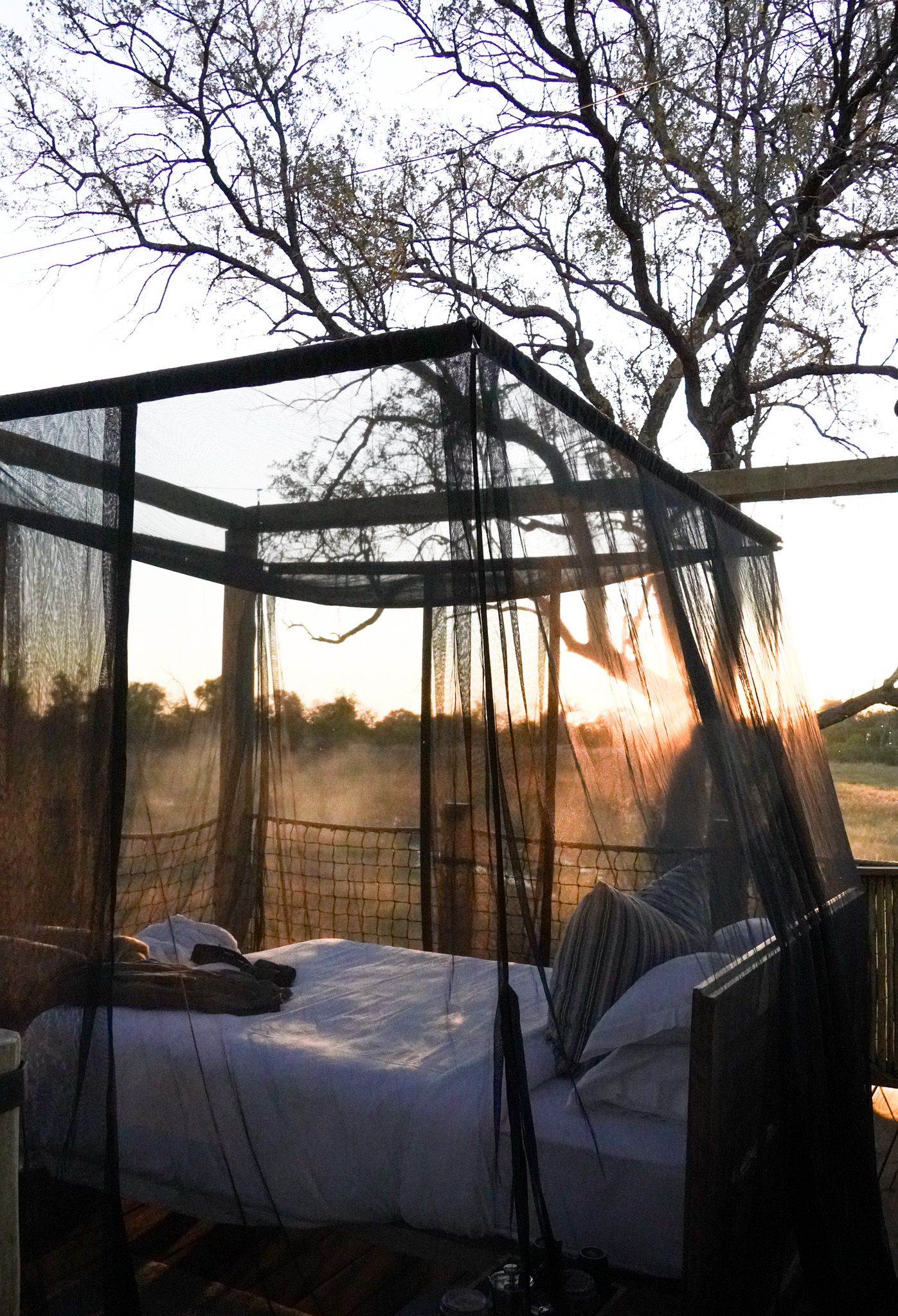 Outdoor canopy bed draped with sheer curtains capturing a magical sunset through trees.