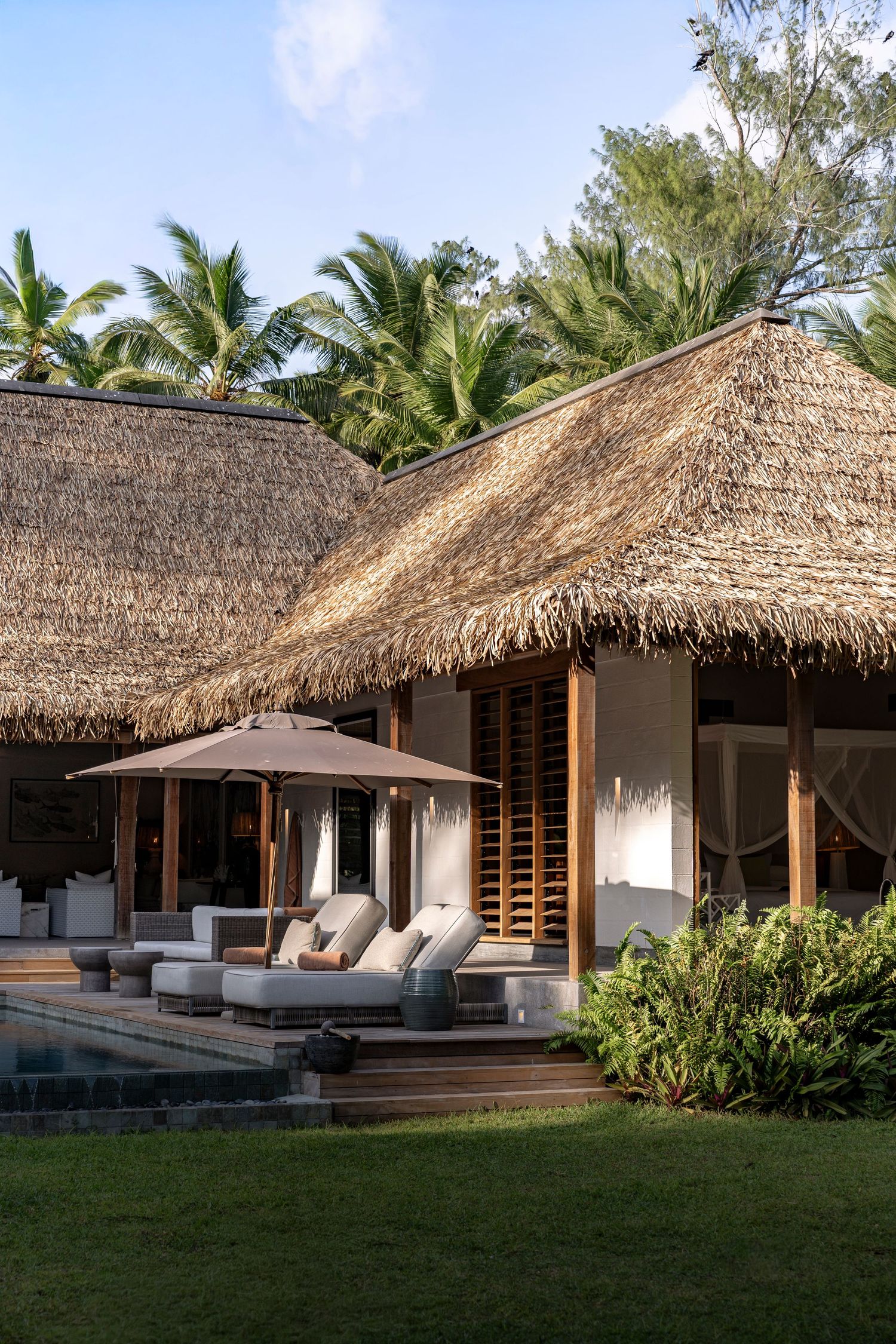 Luxury tropical villa with thatched roof, poolside lounge chairs, umbrella, and lush palm trees in a serene resort setting.