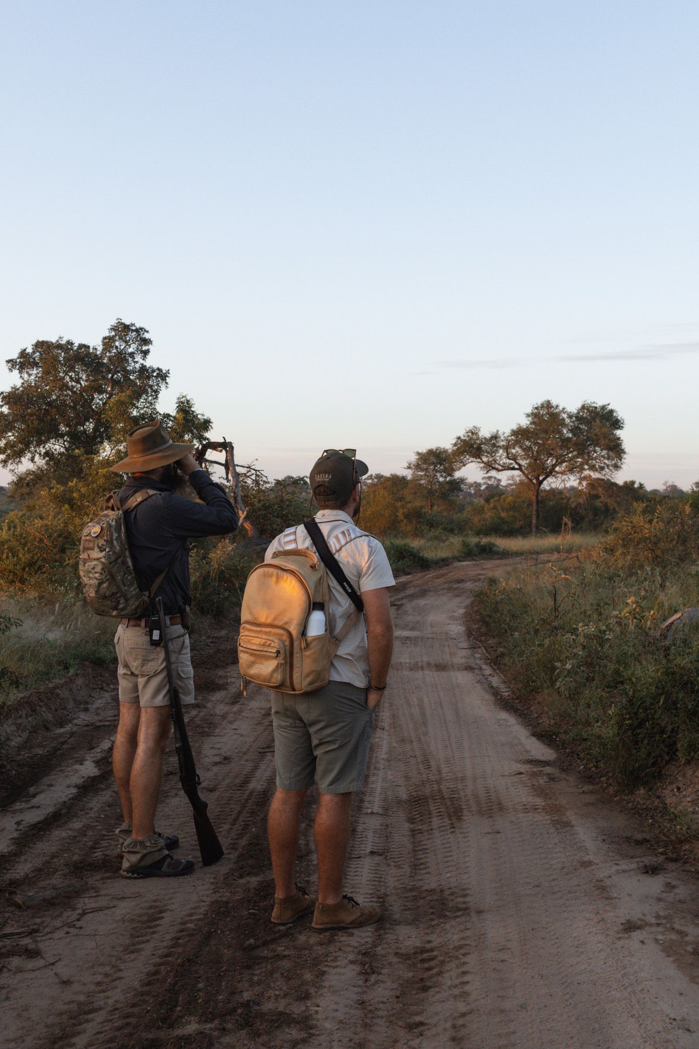 Two photographers with backpacks stand on dirt path observing wildlife.