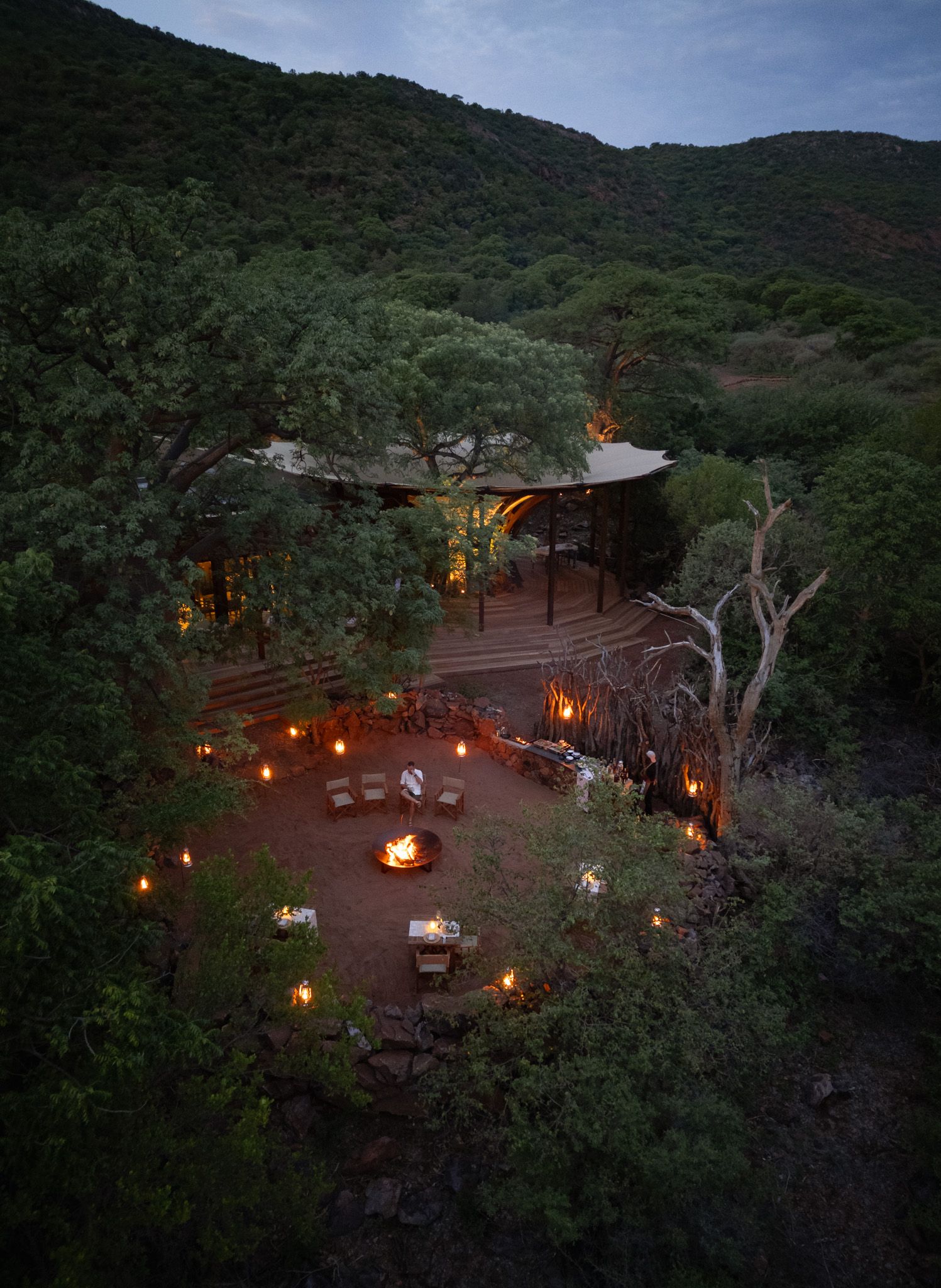 Romantic outdoor dining area lit by lanterns in a forest clearing at dusk.