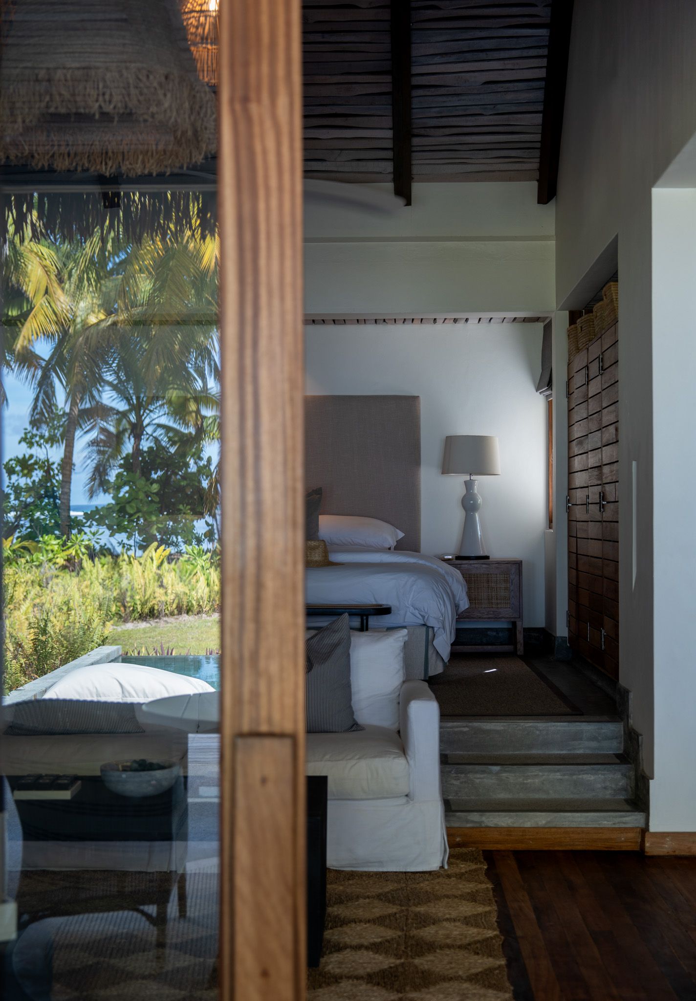 Tropical resort bedroom with white bedding and palm trees visible through large windows.