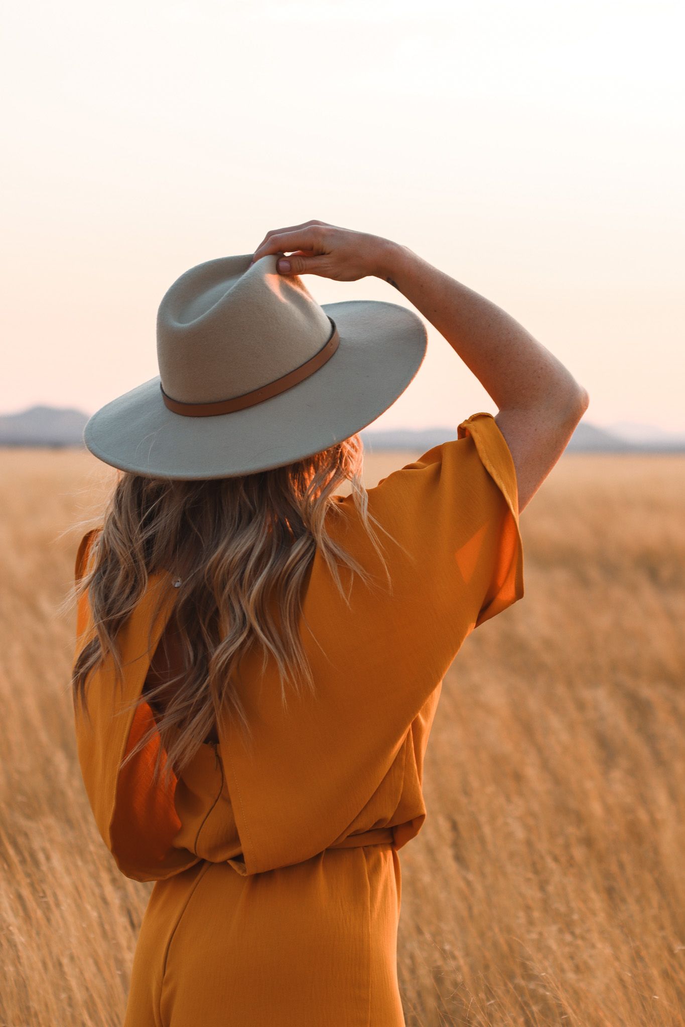 Figure in orange dress and grey hat standing in golden wheat field at sunset.