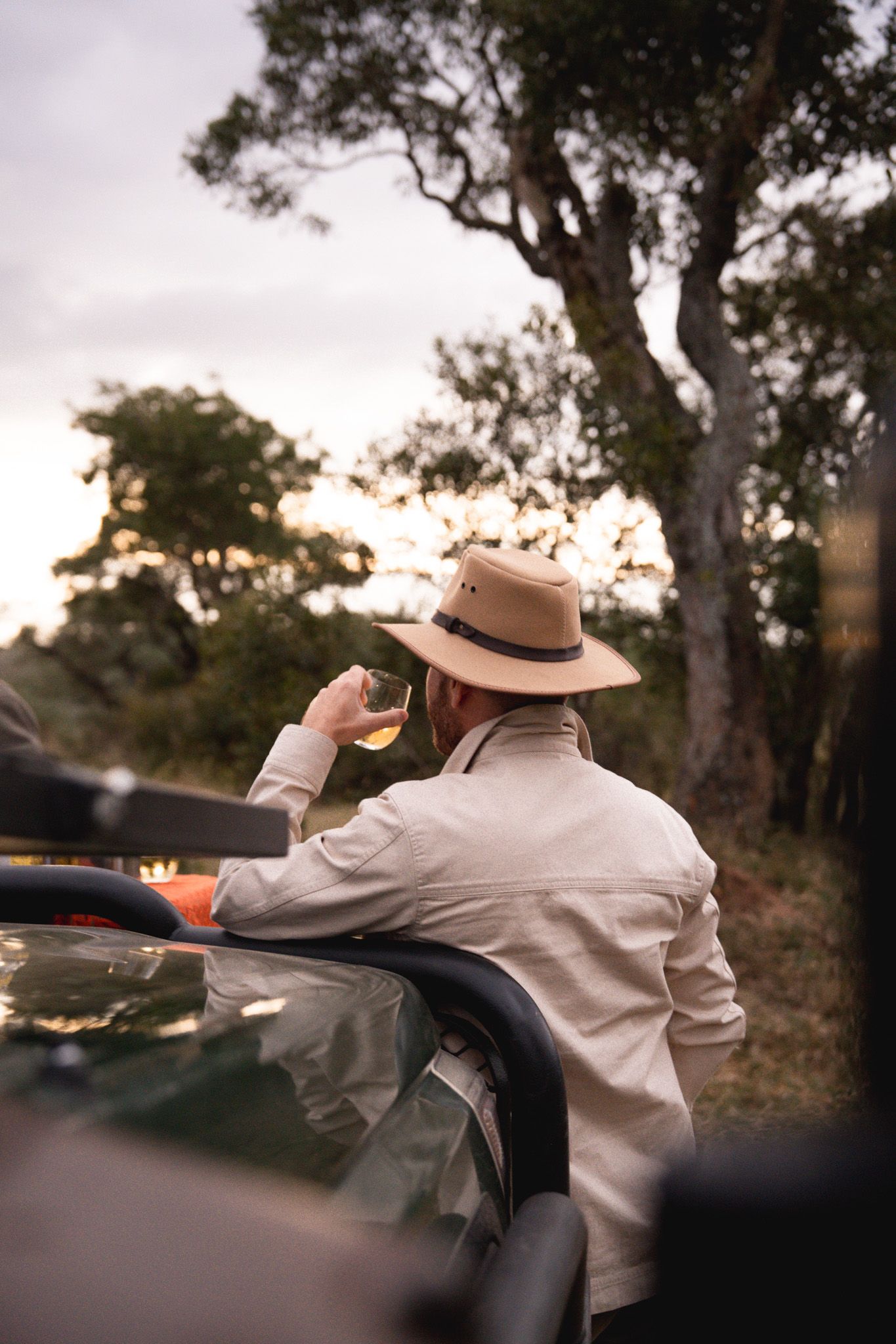 Safari traveler in hat leaning against vehicle while enjoying sunset.
