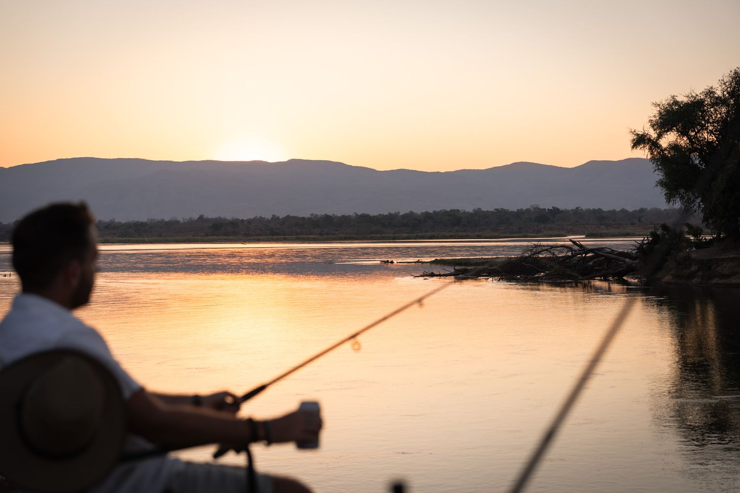 Silhouette of person fishing at sunset on peaceful lake with mountains in background.