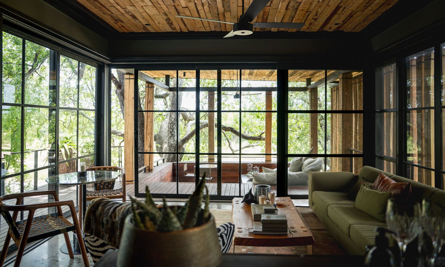Cozy sunroom with wooden ceiling, large windows, green sofa and indoor plants creating a natural ambiance.