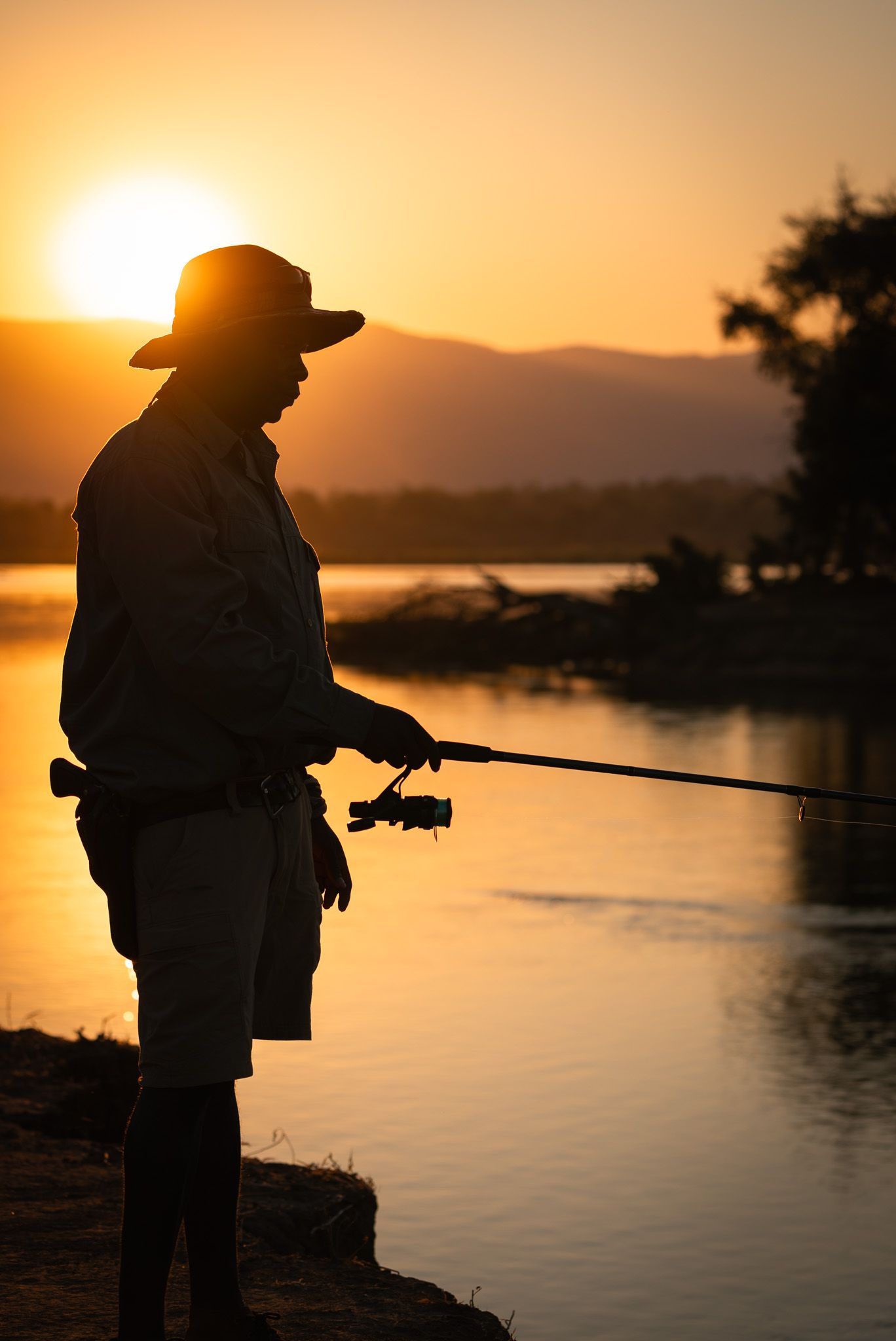 Silhouette of person fishing at sunset by tranquil lake with mountains in background.