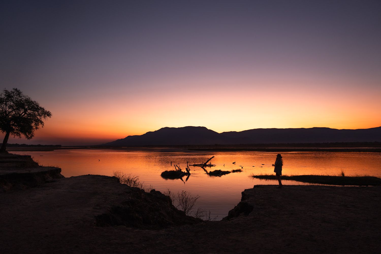 Scenic lake sunset with silhouettes of boats and figure standing on shore against mountain backdrop.