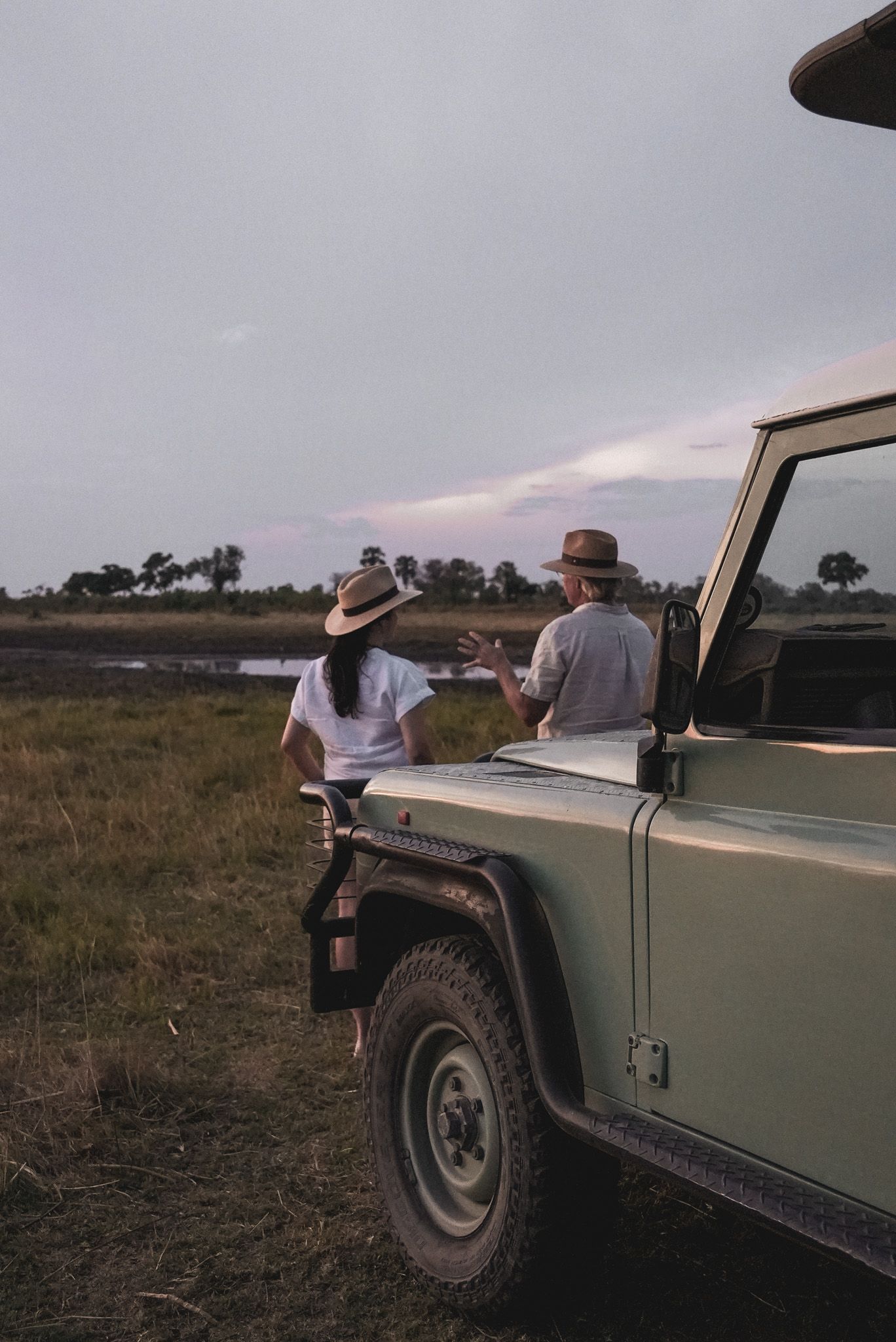 Two people leaning against a vintage Land Rover in an African savanna at dusk.
