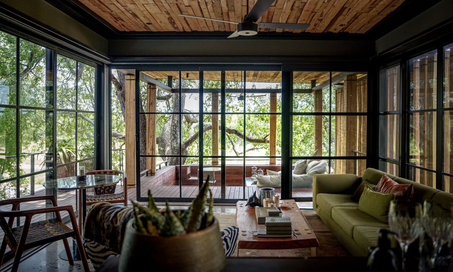 Luxurious treehouse living room with floor-to-ceiling steel windows, green sofa, wood ceiling, and lush forest views.