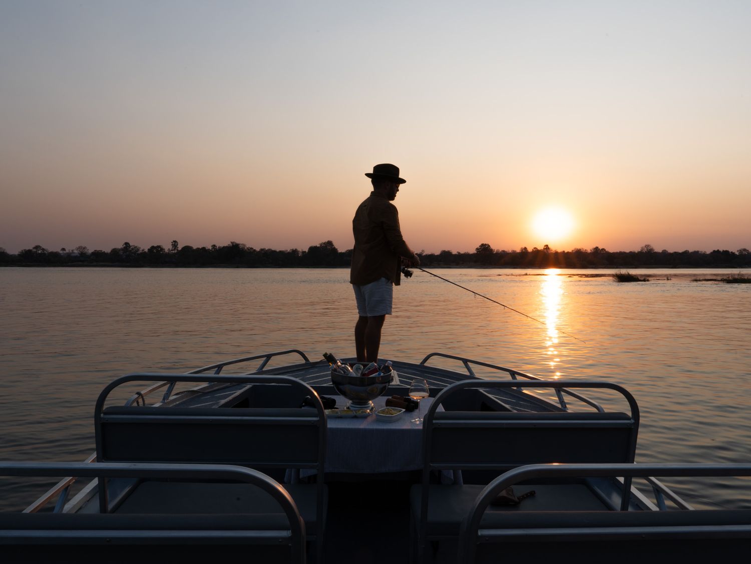 Silhouetted figure fishing from a boat during a golden sunset over calm waters.