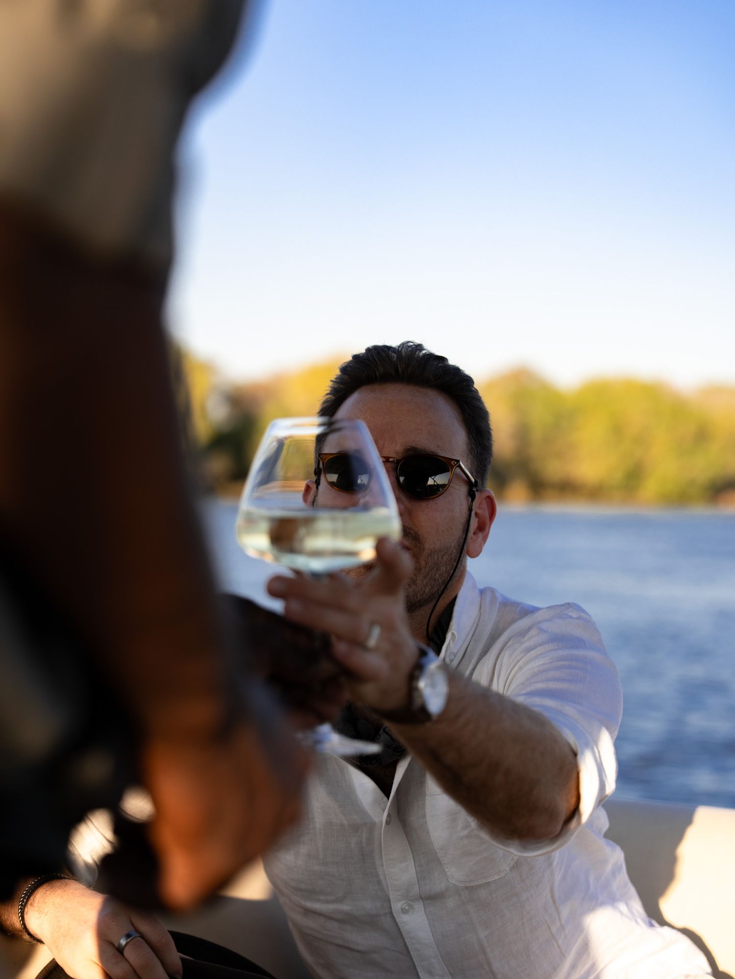 Person holding up a wine glass for a toast against a waterfront sunset backdrop.