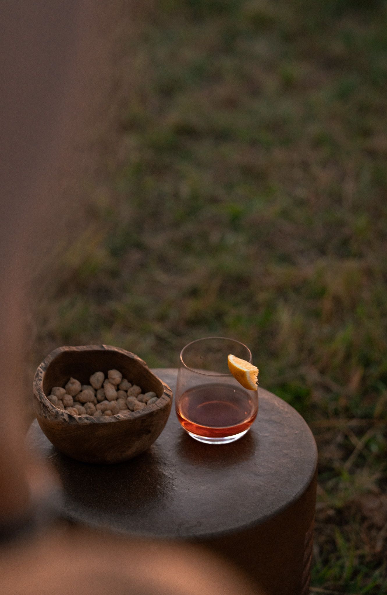 Dark wooden serving board with small bowls of snacks and glass of amber liquid at dusk.