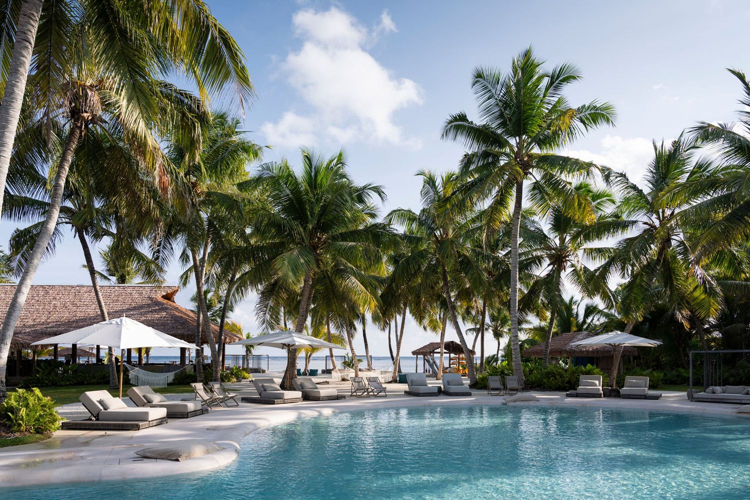 Tropical resort pool surrounded by tall palm trees, lounge chairs, and thatched umbrellas near a serene beachfront.