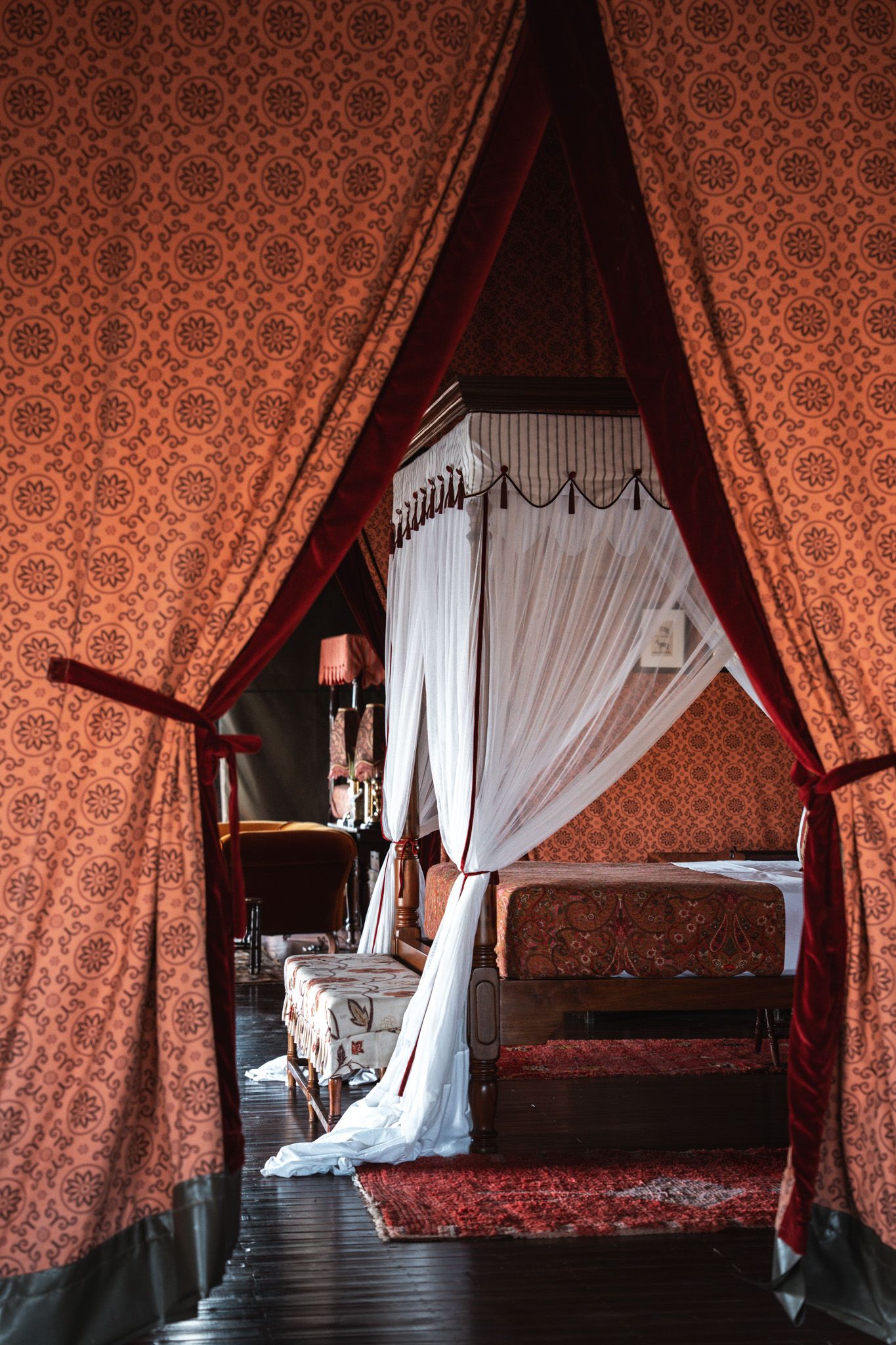 A luxurious canopy bed with red damask curtains and white drapes in a historic bedroom setting.