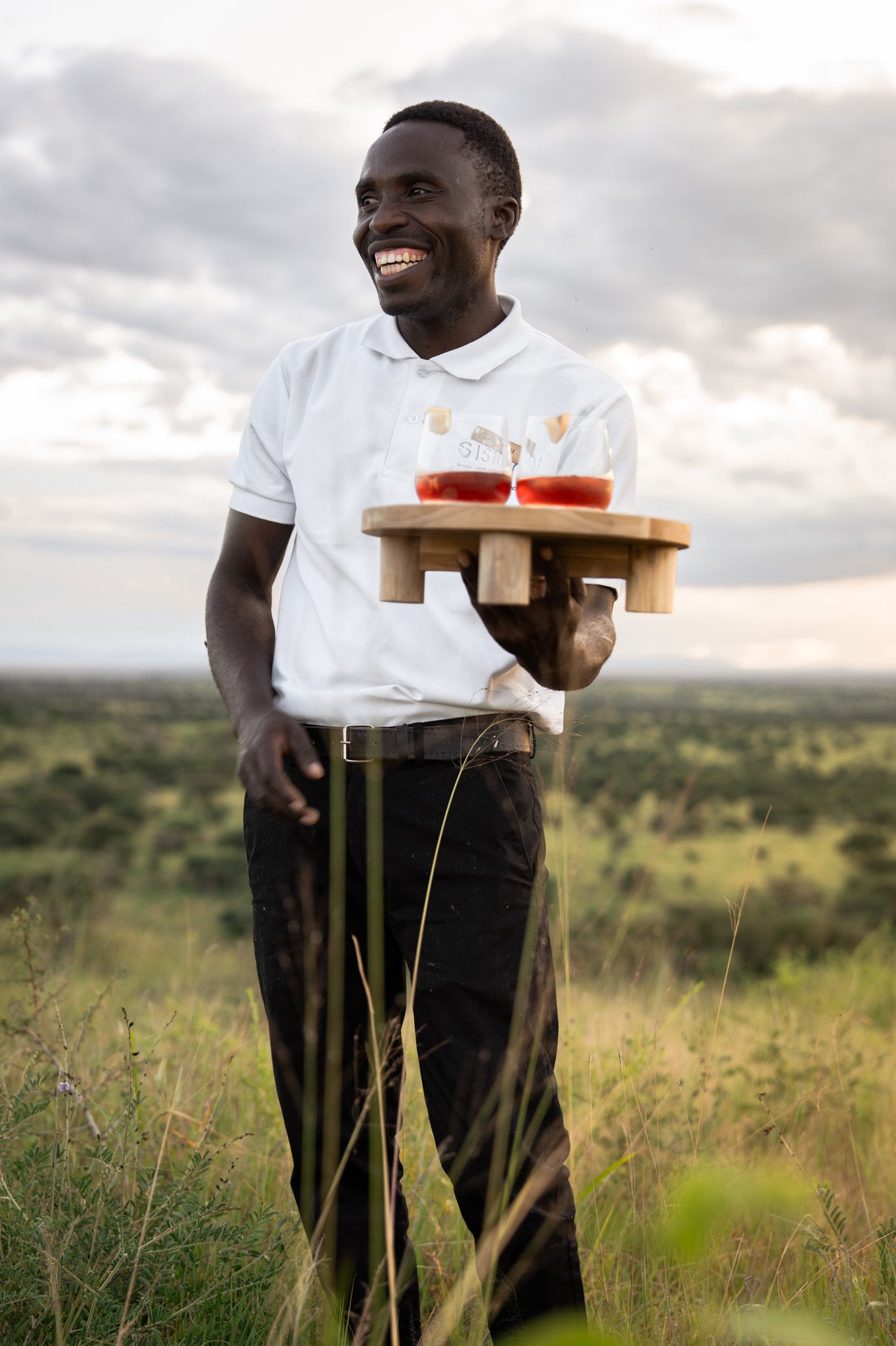 Staff member carrying wooden serving tray across grassy savanna landscape.