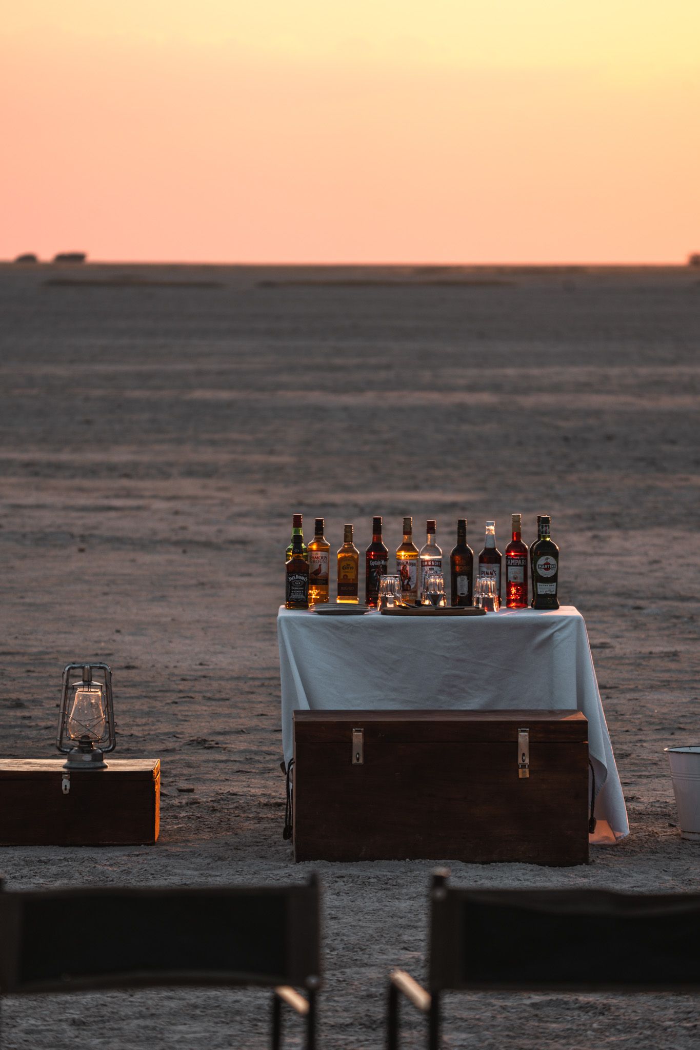 Bar setup with various liquor bottles on tables against a sunset sky.