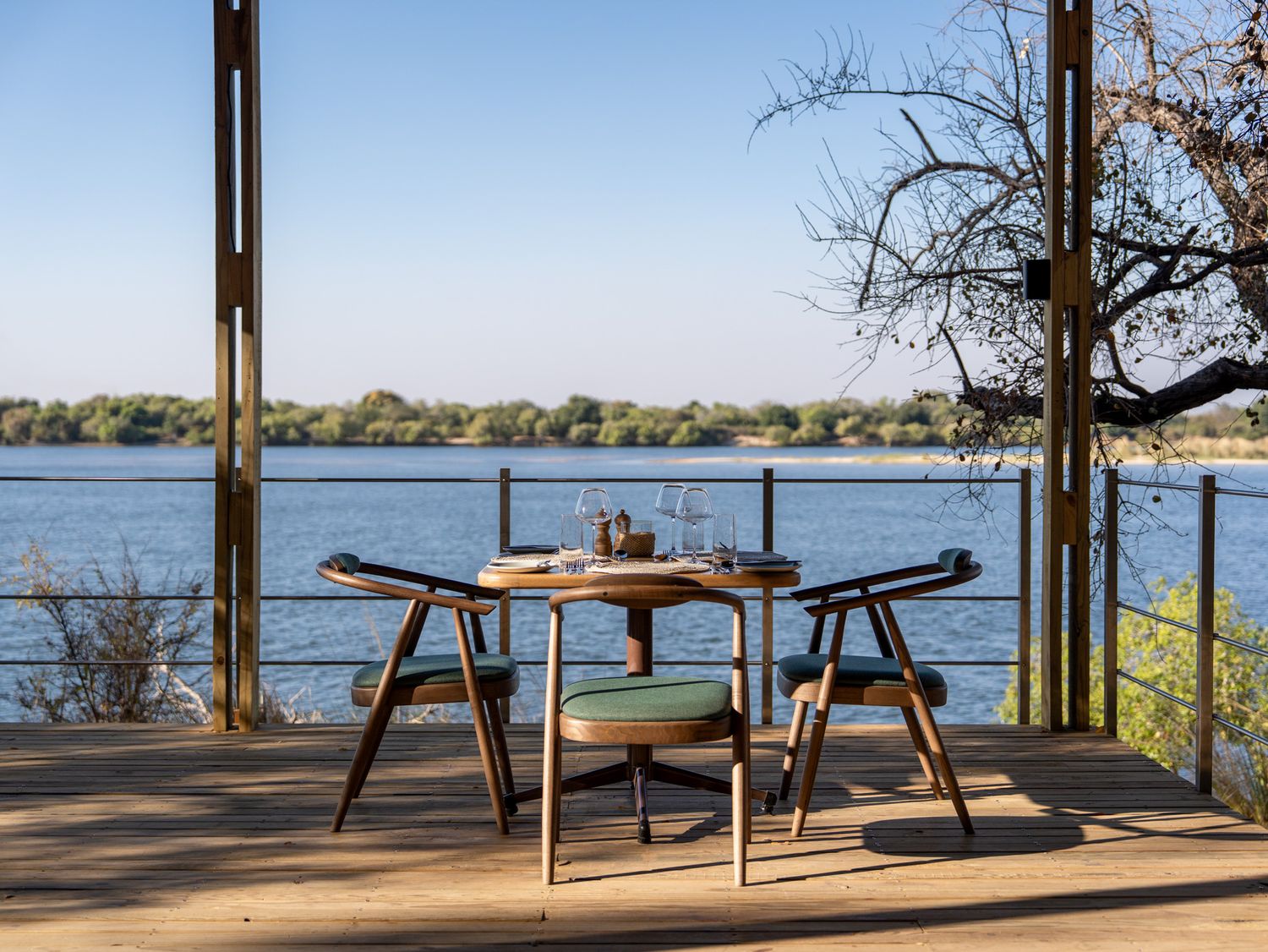 A scenic lakeside dining setup with a small table and chairs on a wooden deck overlooking calm waters.