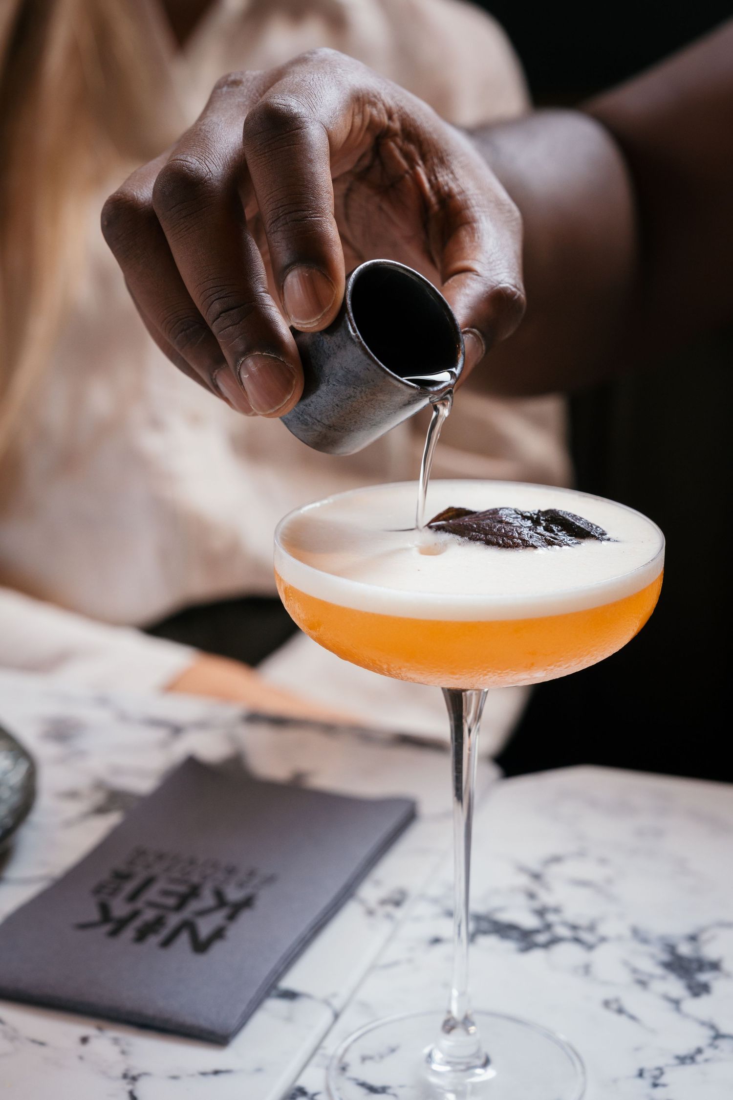 Bartender pouring a finishing touch into an orange cocktail on a marble surface.