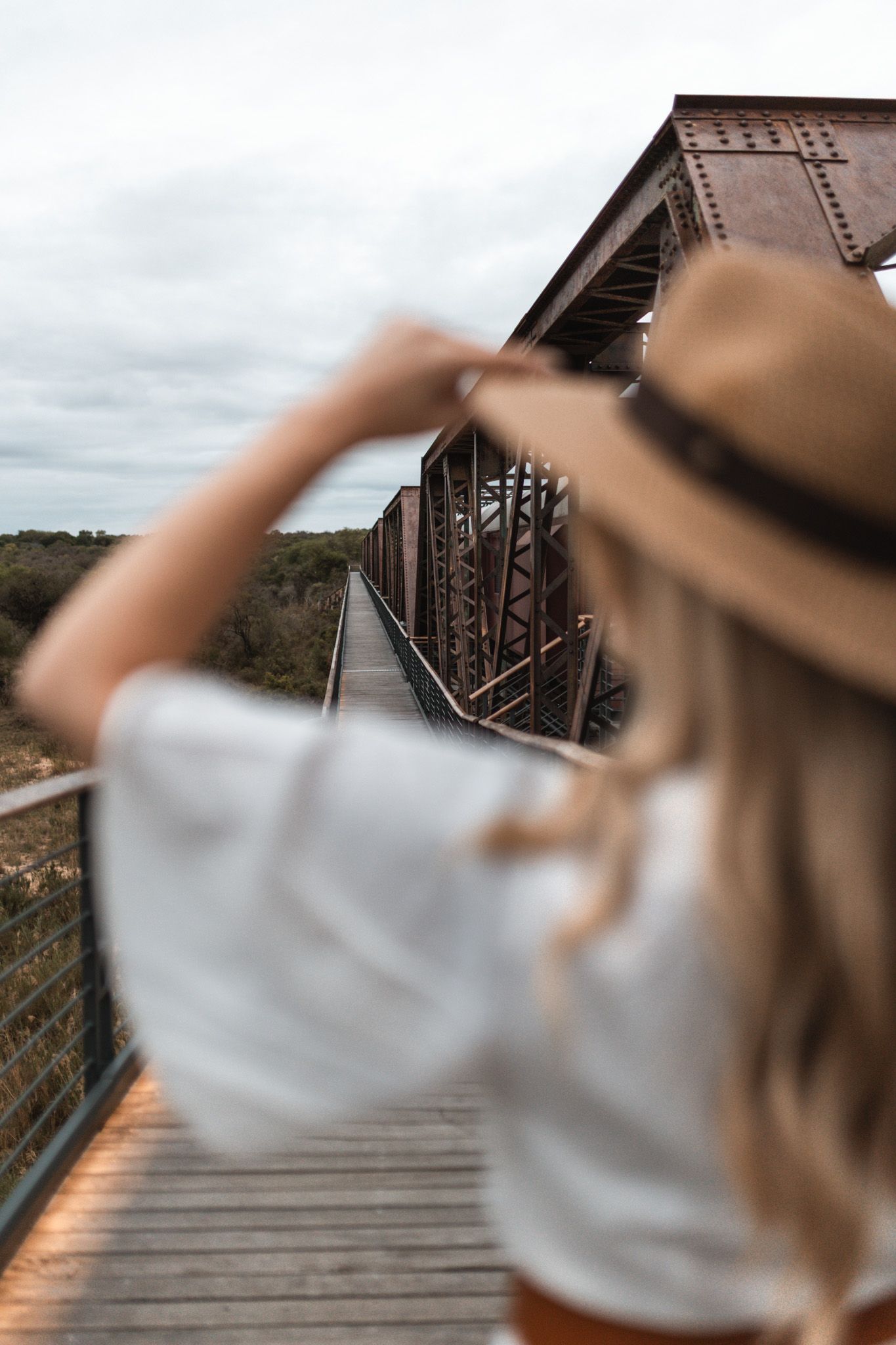 Person wearing hat looks out over rustic wooden bridge in scenic landscape.