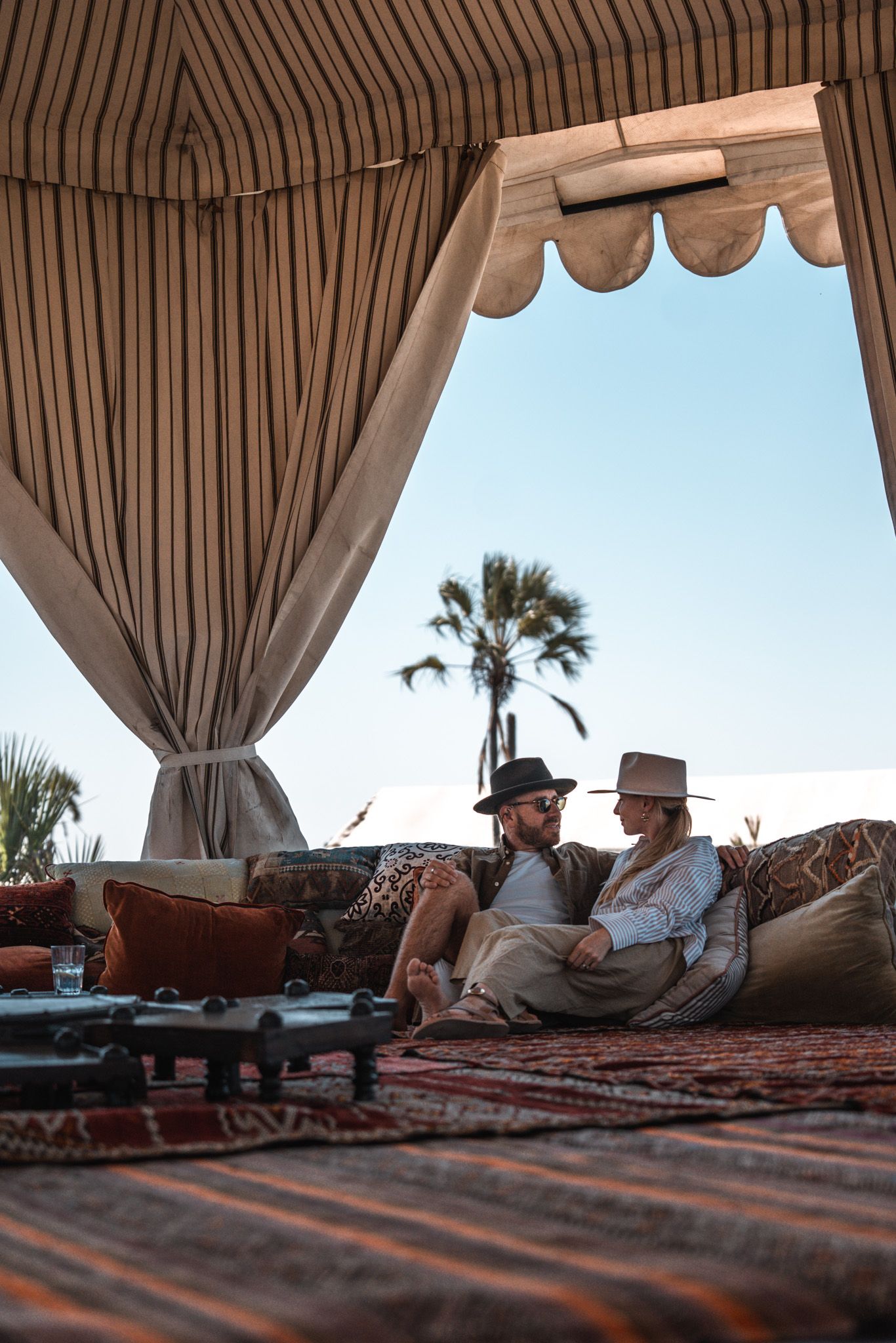 Two people lounging on cushions inside a striped safari tent with palm trees visible outside.
