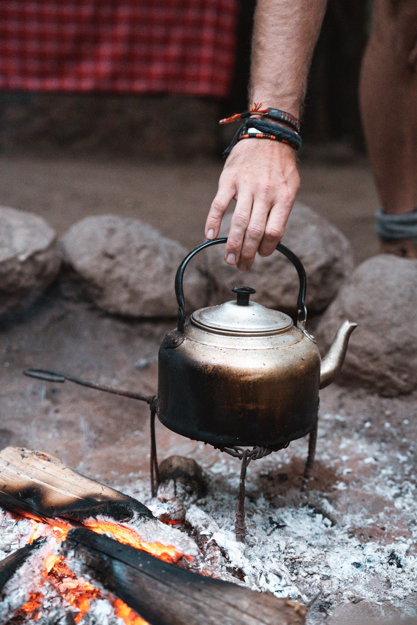 Kettle heating over hot coals on rustic campfire with red checkered cloth in background.