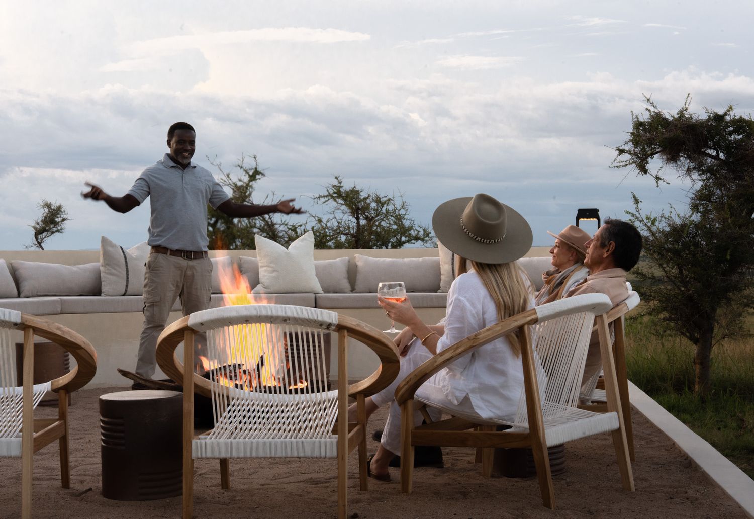 Guests gather around outdoor fire pit on luxury safari lodge deck at dusk.