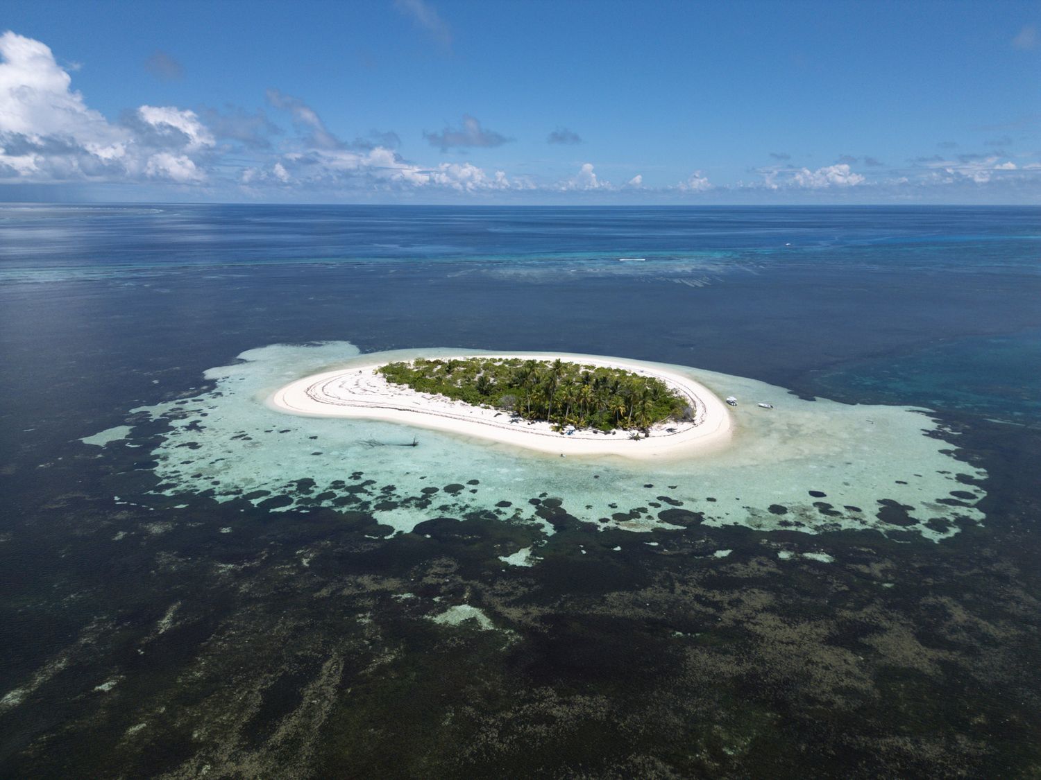 Small circular tropical island surrounded by coral reef and turquoise ocean waters.