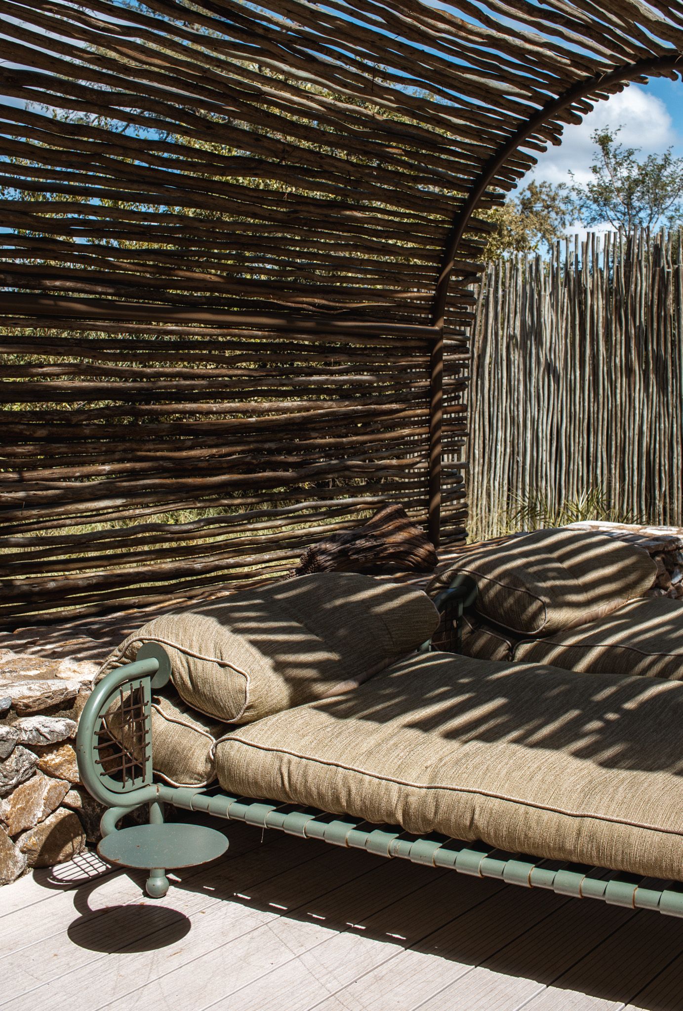 Outdoor daybed with beige cushions under rustic thatched roof creating dramatic shadows.