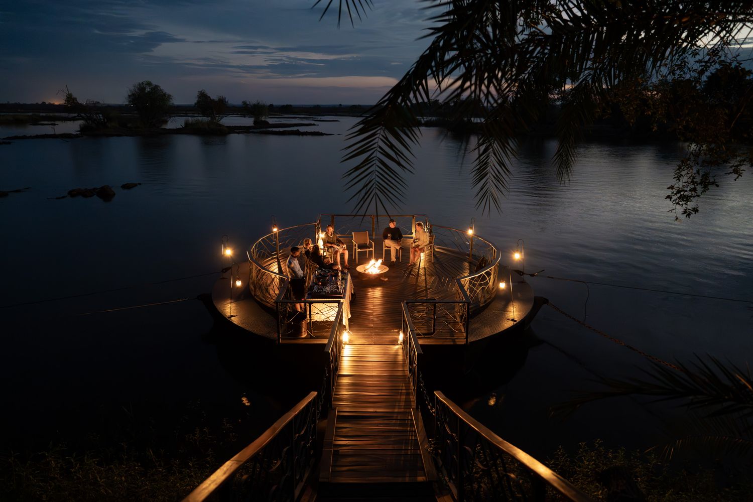 Illuminated circular deck with candles over tranquil water at dusk, connected by a lit wooden walkway.