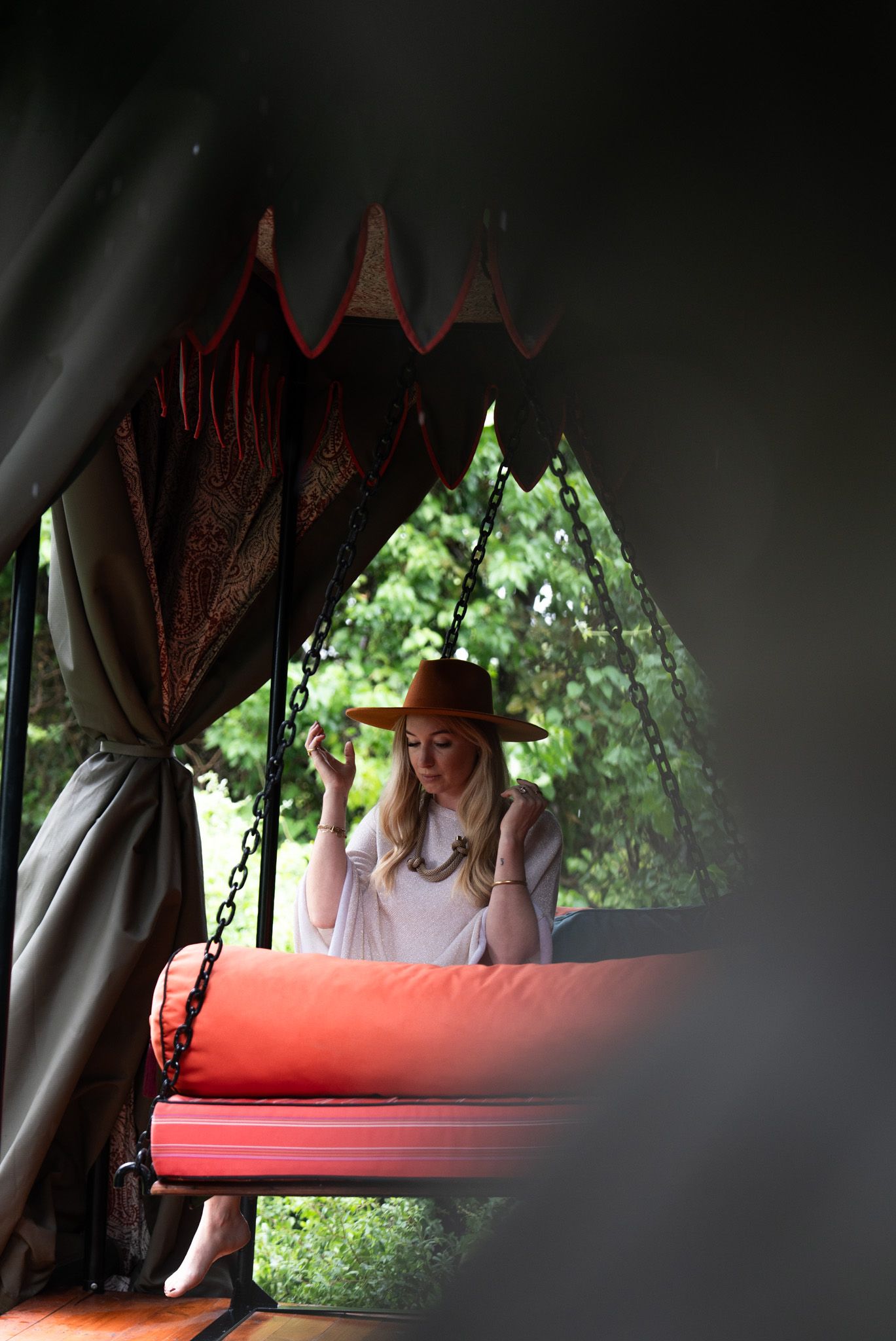 A person wearing a sun hat sits on a red cushioned bench under brown curtains in a peaceful outdoor setting.