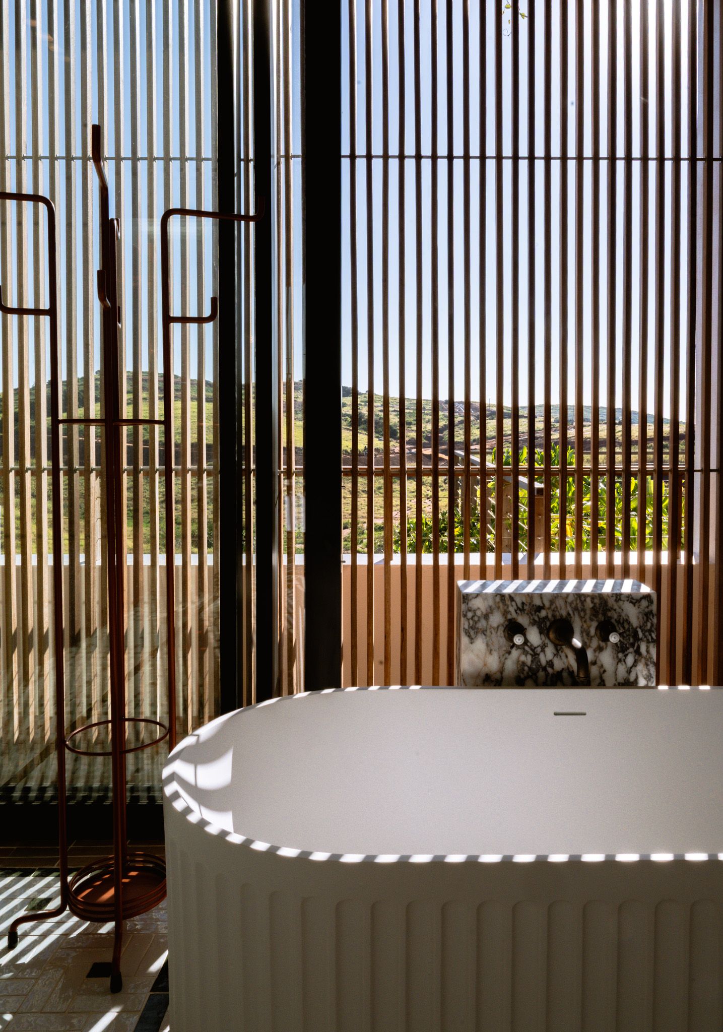 Freestanding bathtub near vertical blinds creating dramatic light patterns.
