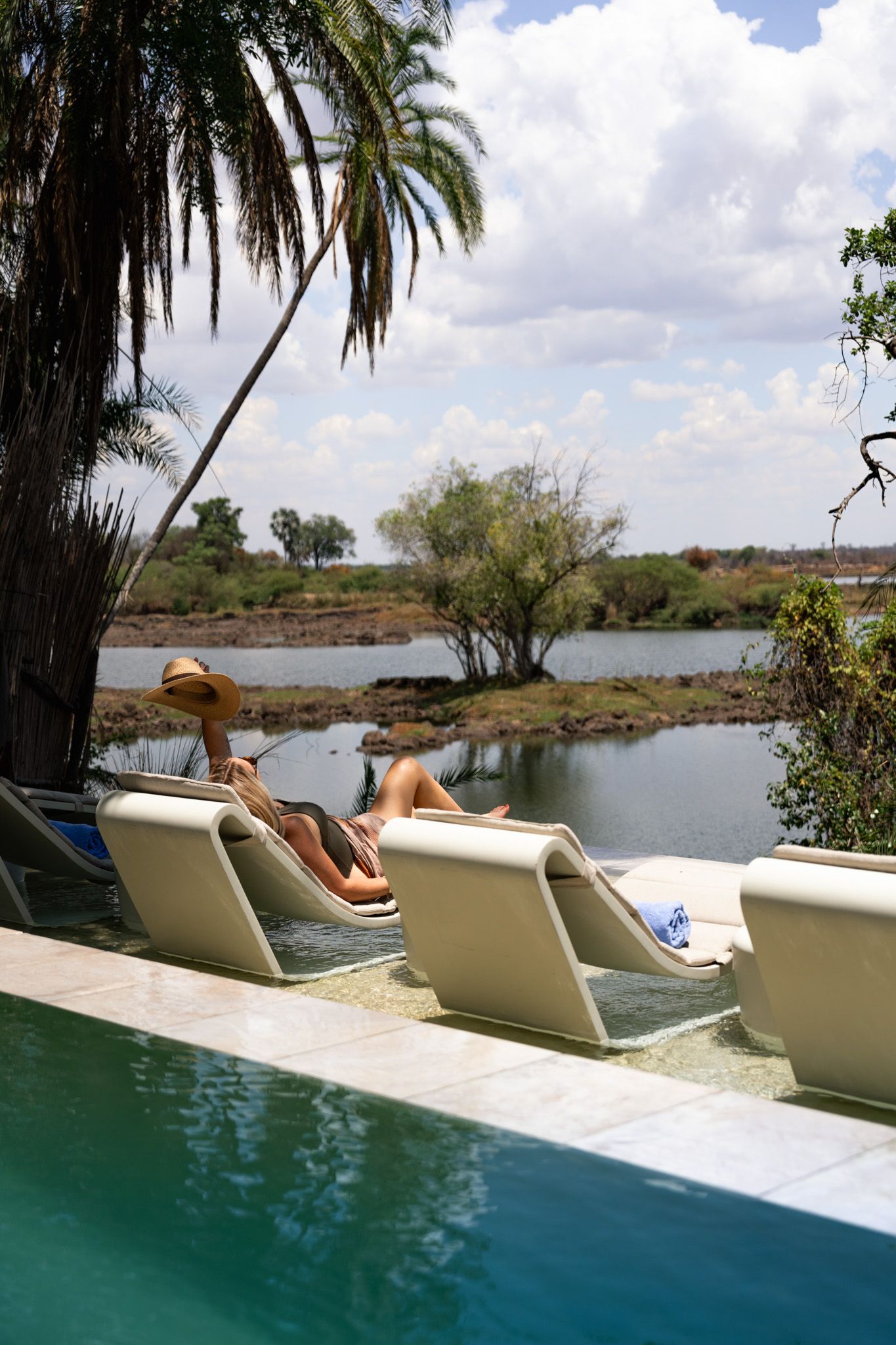 Poolside loungers overlooking a tranquil lake with palm trees and natural landscape.