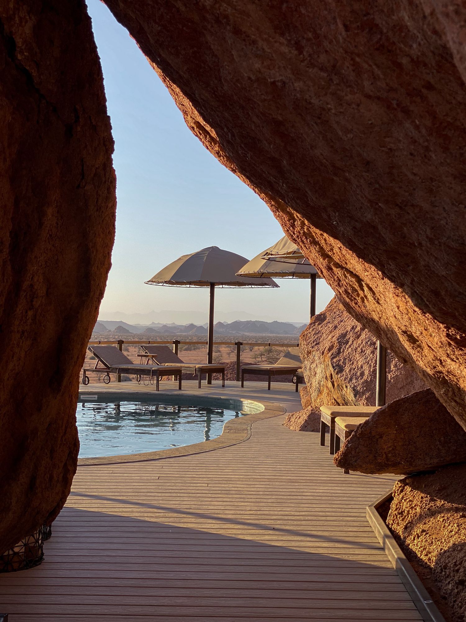 Swimming pool with umbrella viewed through rock formation cave opening in desert landscape.