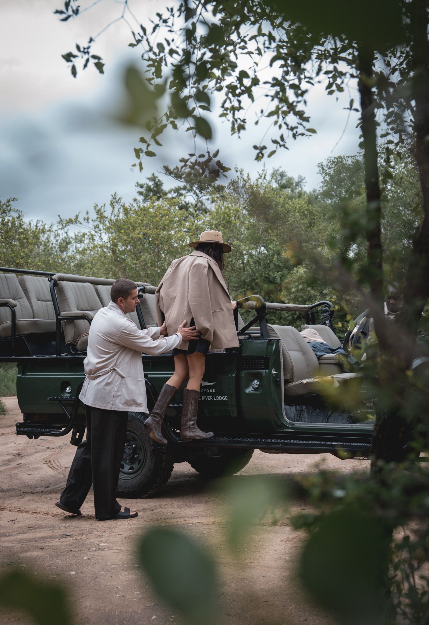 People work on a vintage green Land Rover parked on a dirt road surrounded by foliage.