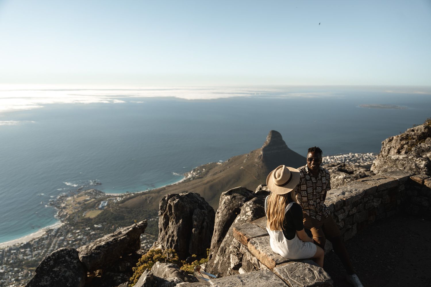 Two tourists admiring the coastal view from a rocky mountain overlook with dramatic ocean scenery.