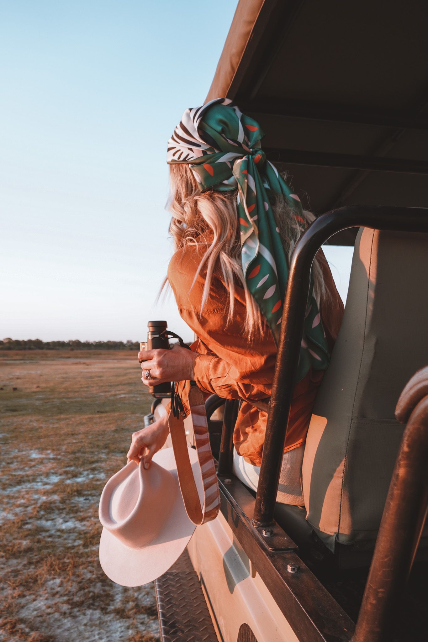 An adventurer in orange attire leans out of a safari vehicle against a clear blue sky.