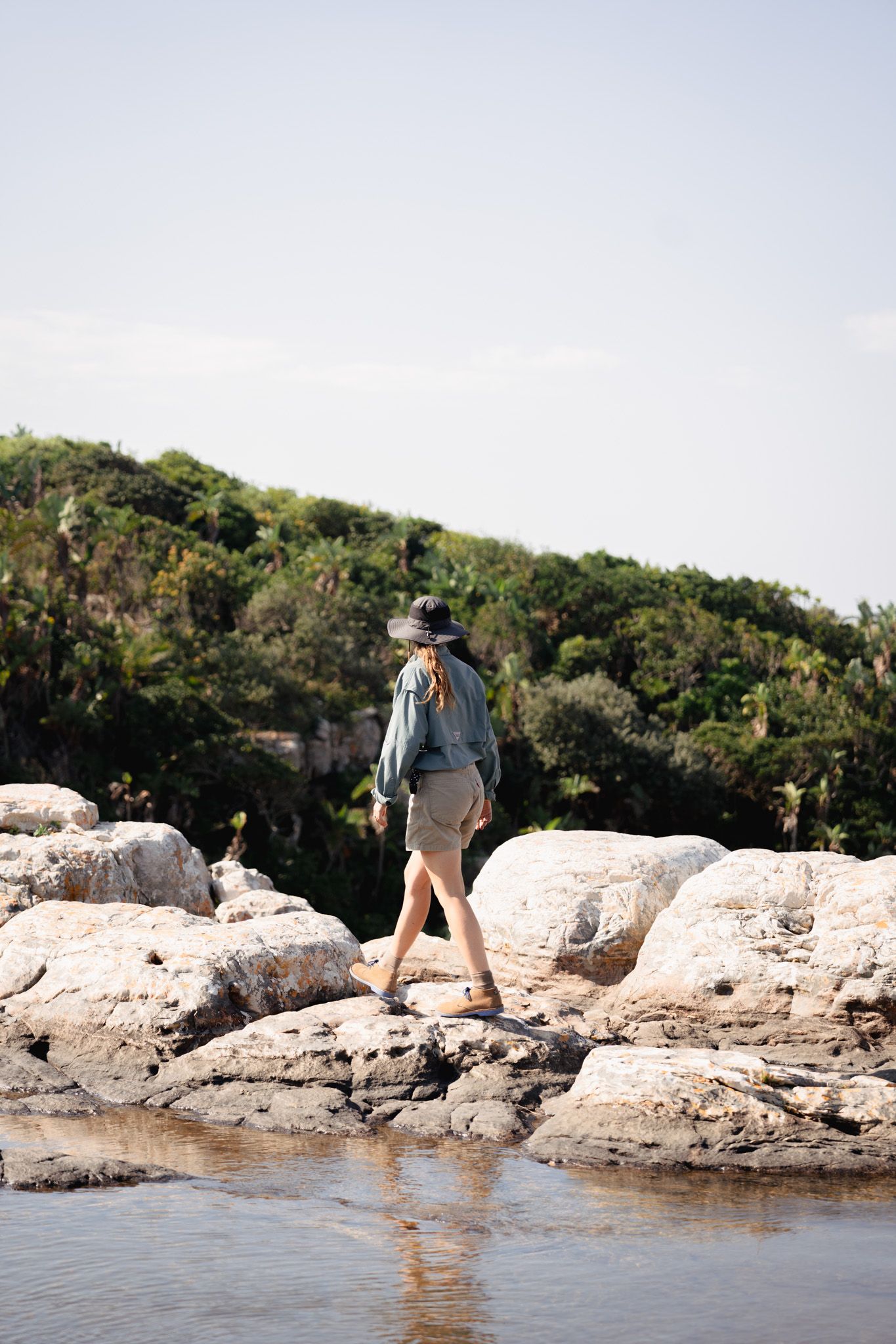Figure in hat walking along rocky coastline with lush vegetation in background.
