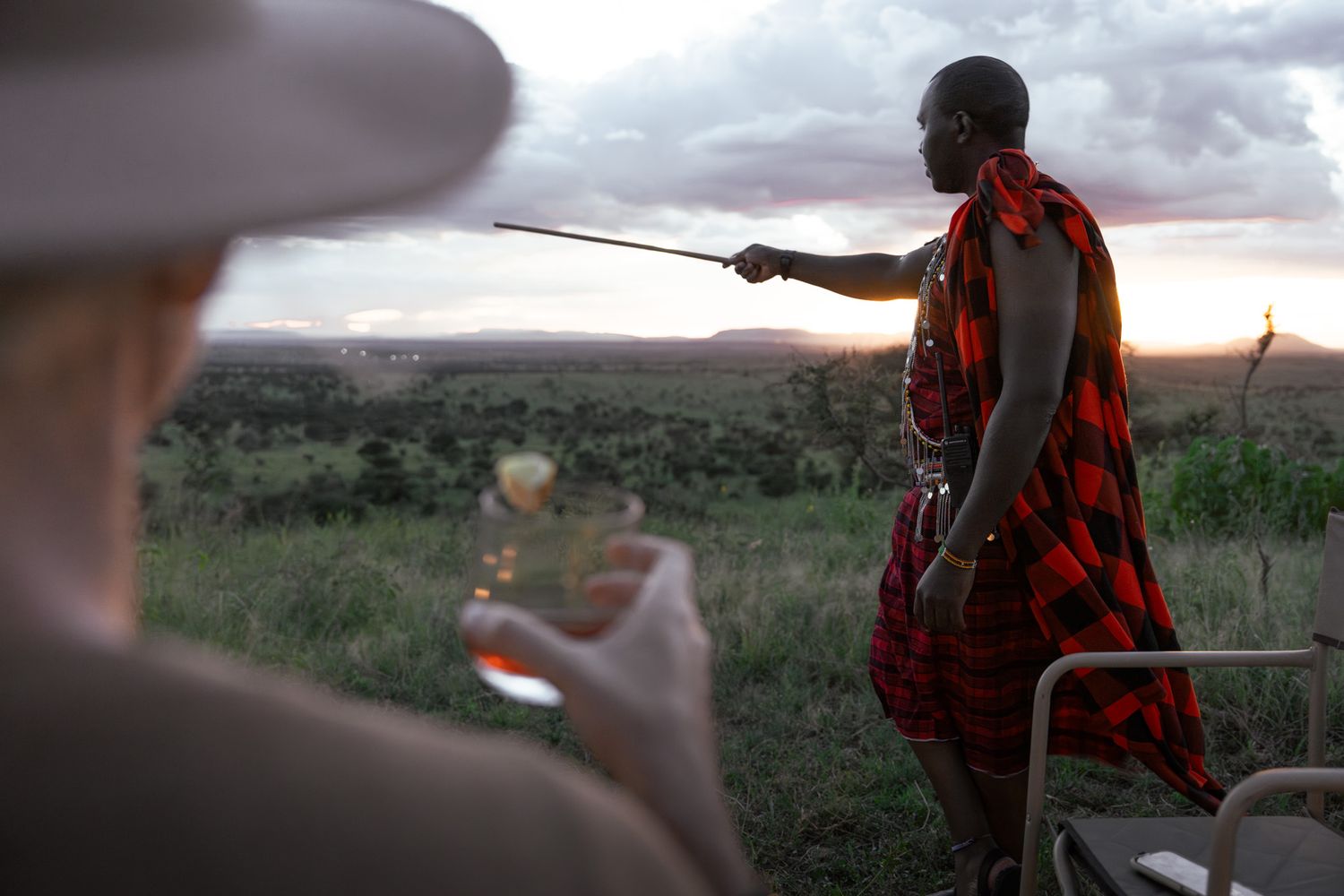 Masai guide in traditional red clothing points across savanna landscape at sunset while safari guest observes.