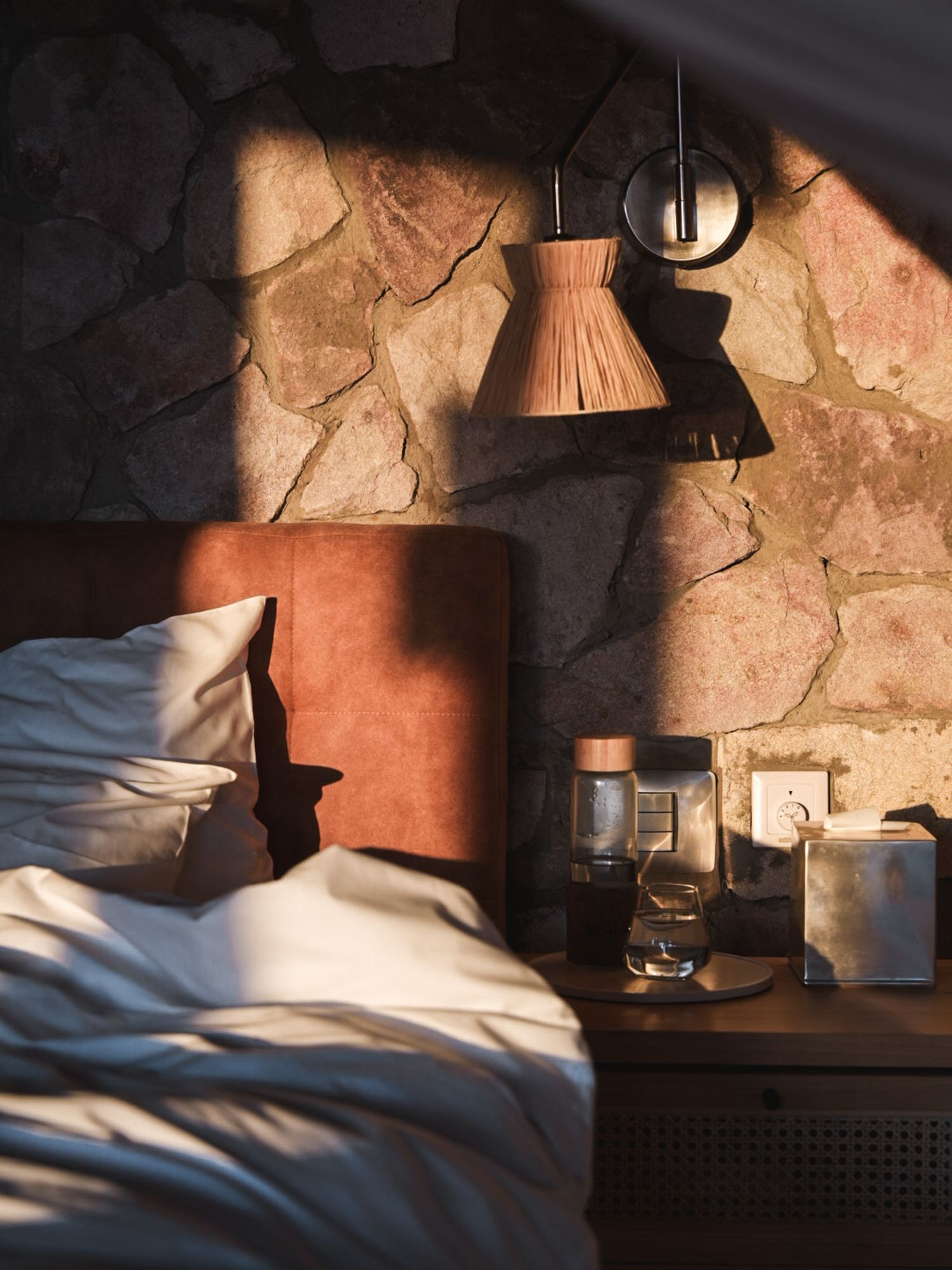 Cozy bedroom corner with stone wall, pendant lamp, and white bedding in dramatic lighting.