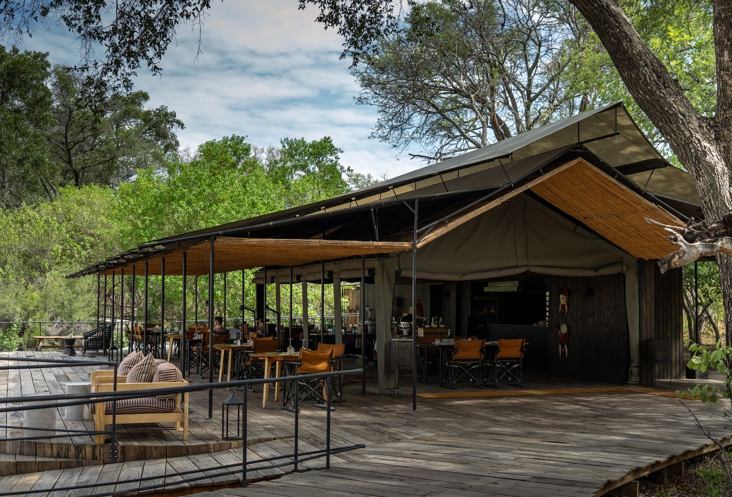 Modern safari lodge with wooden deck and covered dining area surrounded by lush greenery.