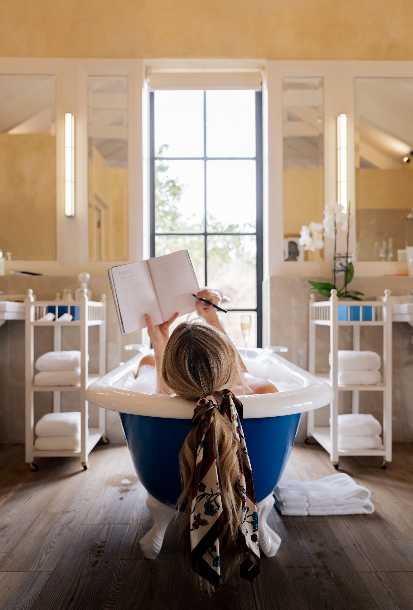 Reading in a luxurious blue bathtub surrounded by white decor and natural light from tall window.