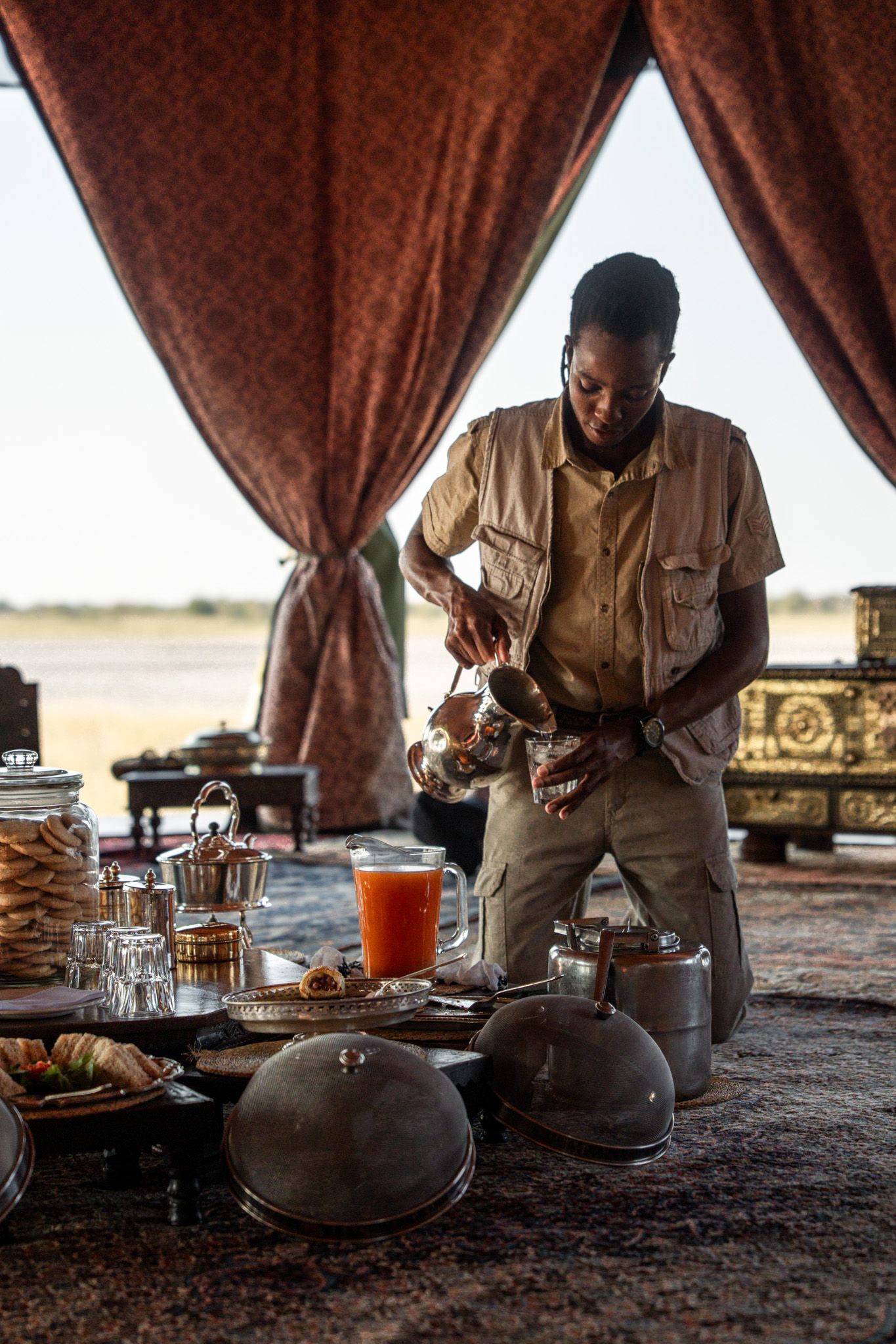 Safari camp dining setup with person preparing refreshments at elegant table setting.