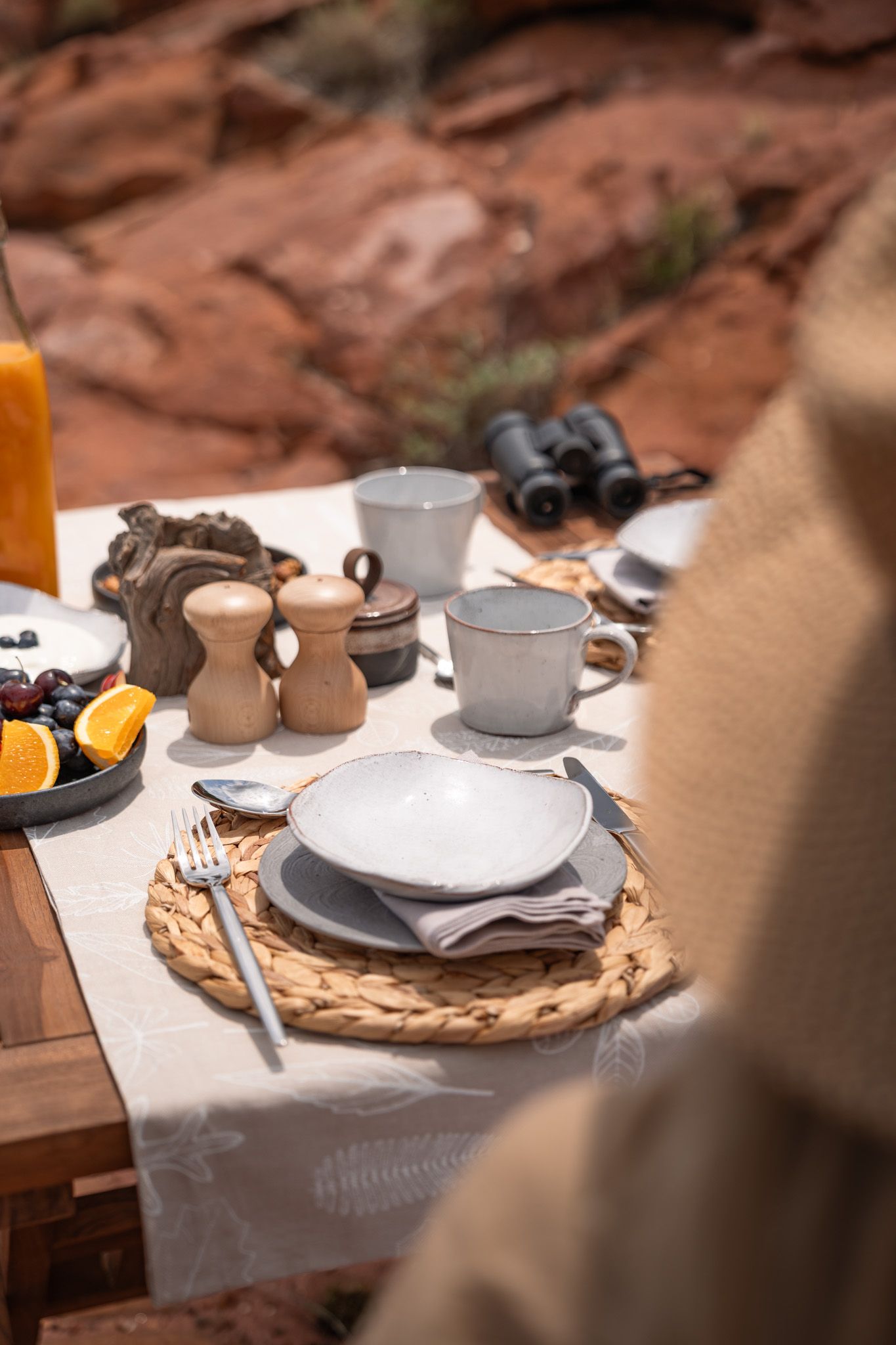 Rustic outdoor table setting with ceramics, teacups and woven placemats against red rock backdrop.
