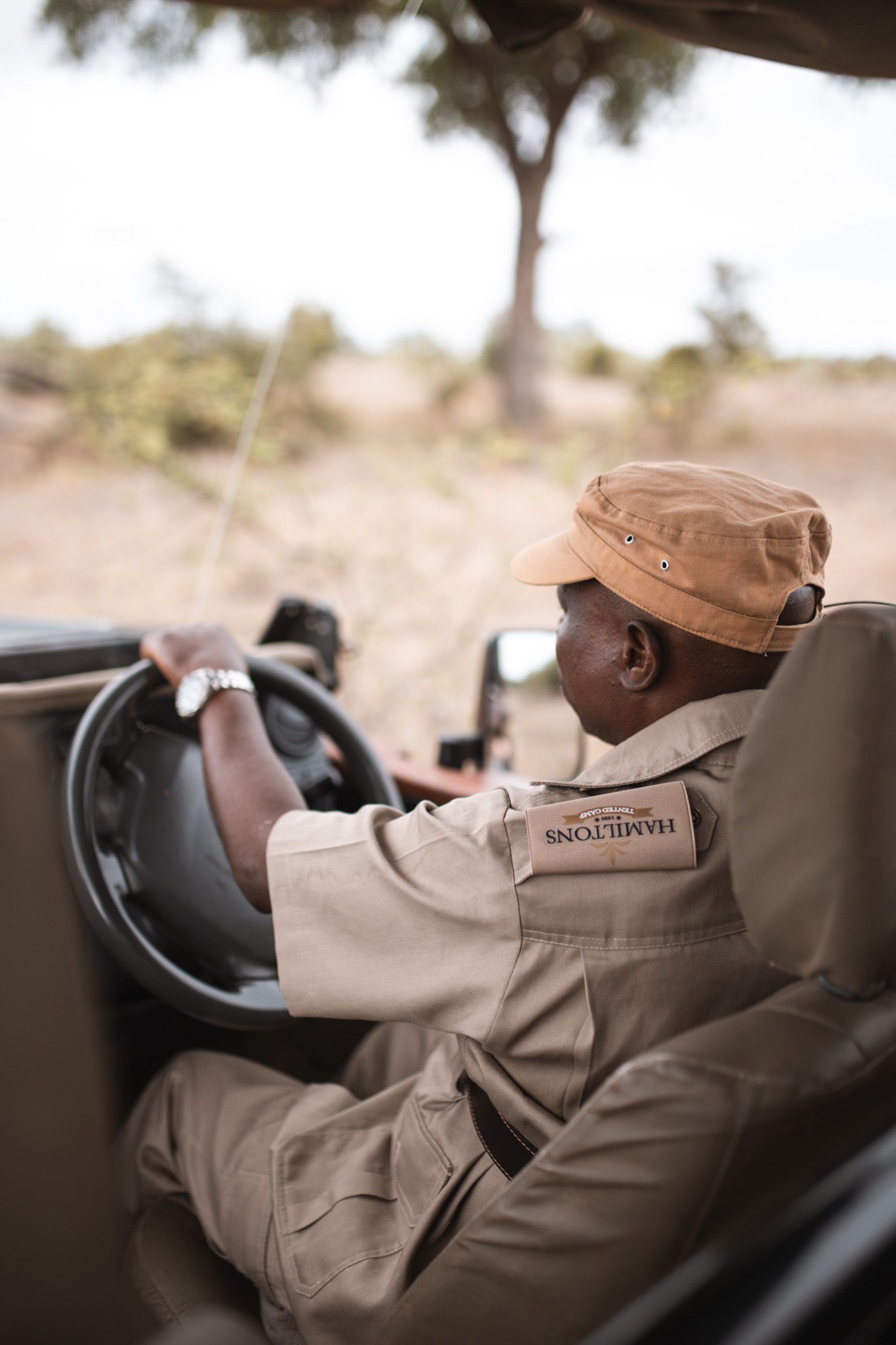 Safari guide in khaki uniform and cap driving a safari vehicle through wilderness terrain.