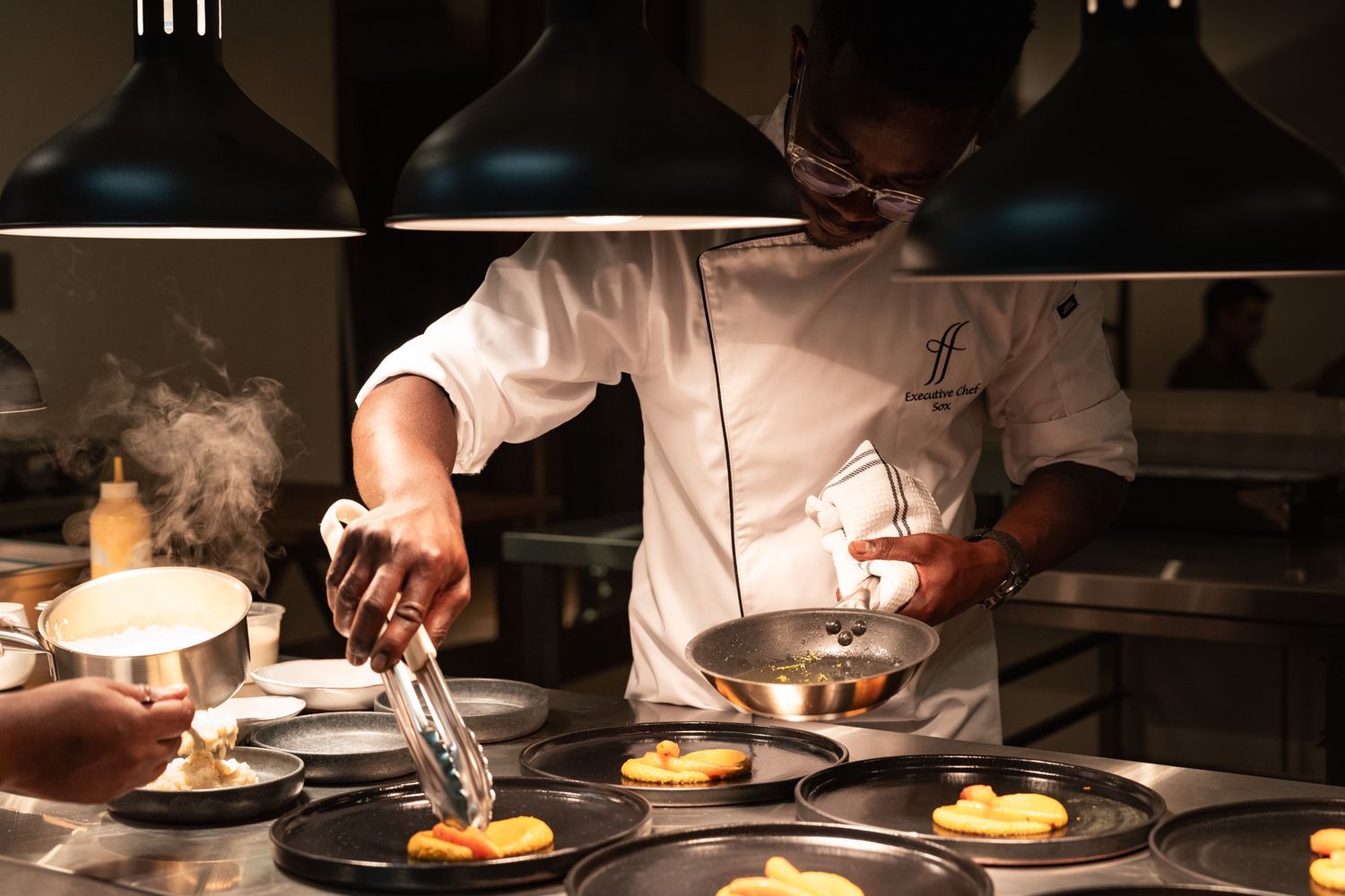 Chef plating multiple dishes in restaurant kitchen under pendant lights.