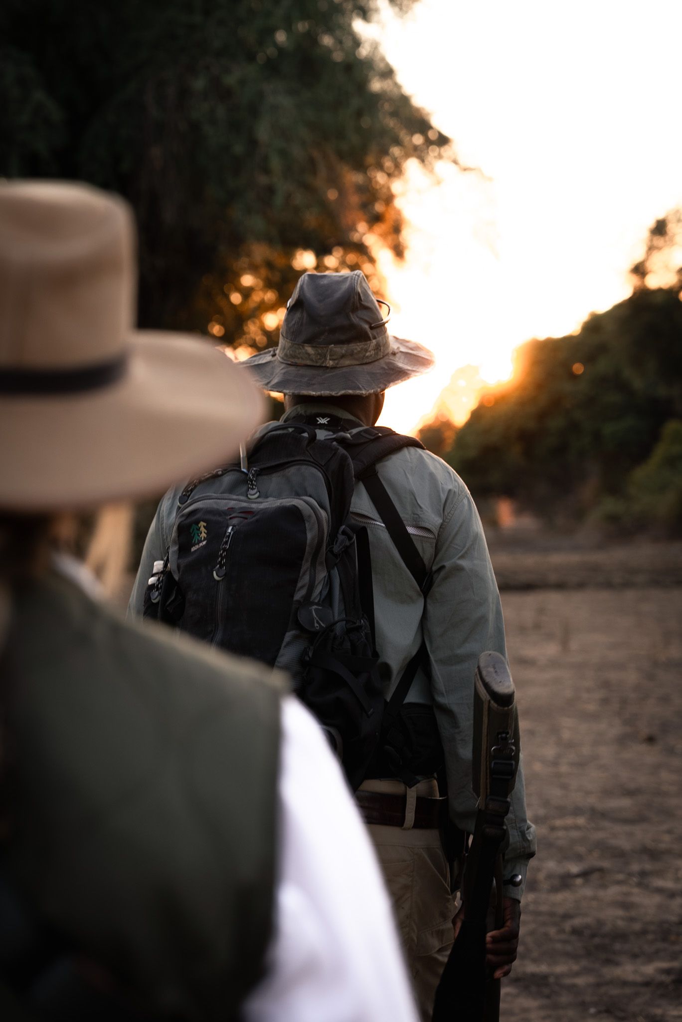 Safari guides walking along trail at sunset with backpacks and wide-brimmed hats.
